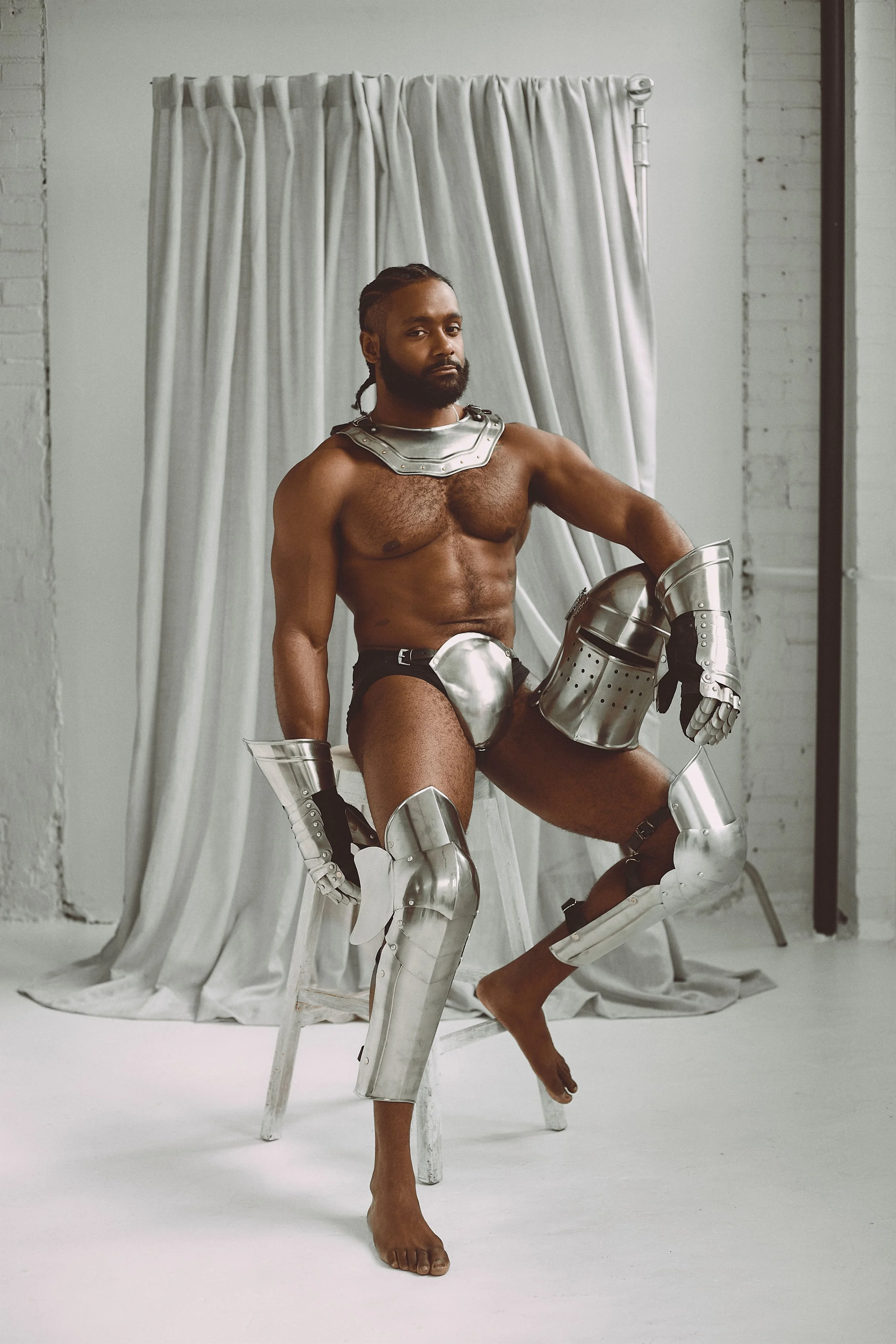 A man with a beard and braided hair posing shirtless on a white chair, wearing metallic armor pieces on his arms, legs, neck, and waist, in front of a white backdrop with white curtains.