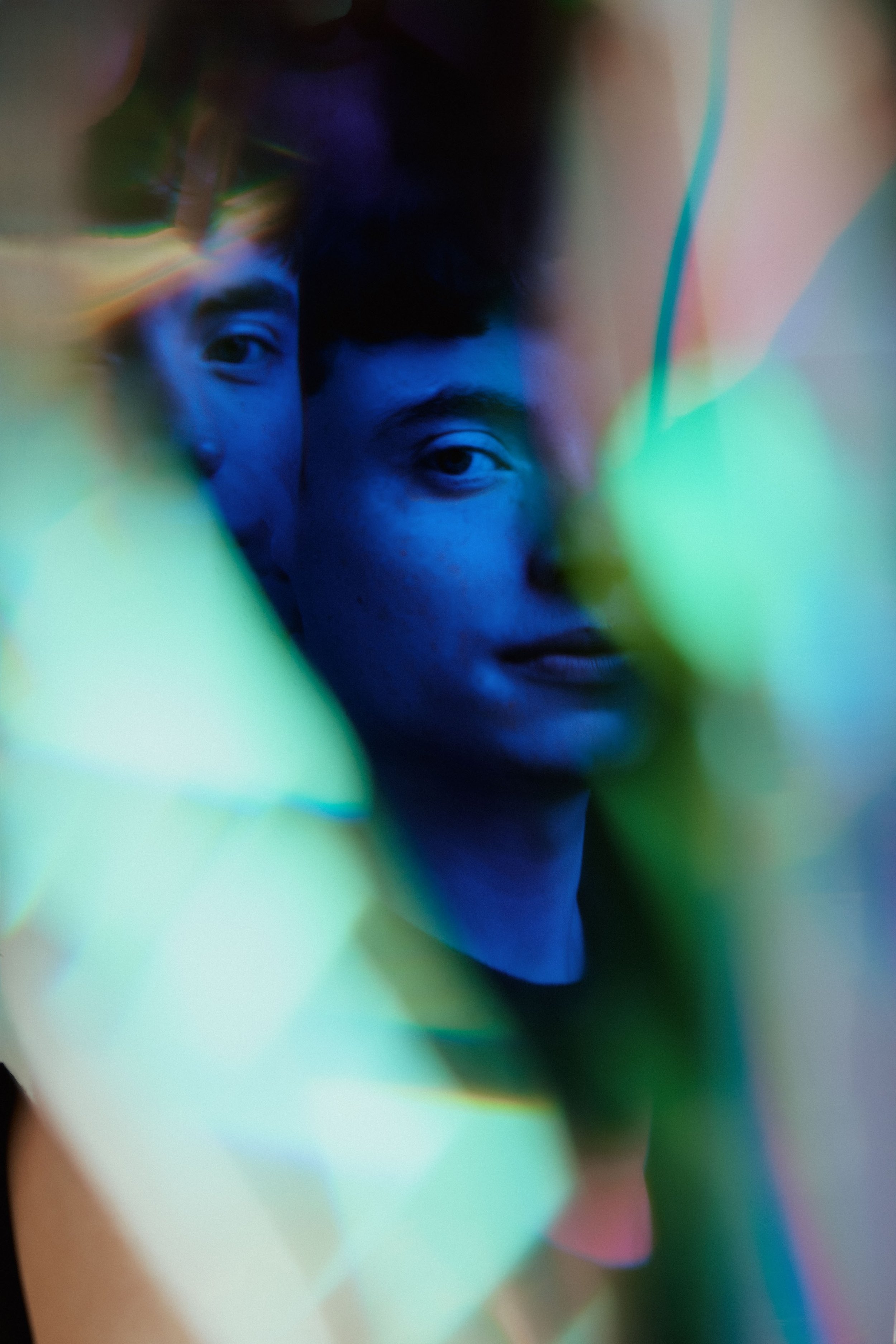 A close-up photo of a person's face with a mix of dark and illuminated areas, surrounded by colorful, abstract light streaks in green, blue, and pink.