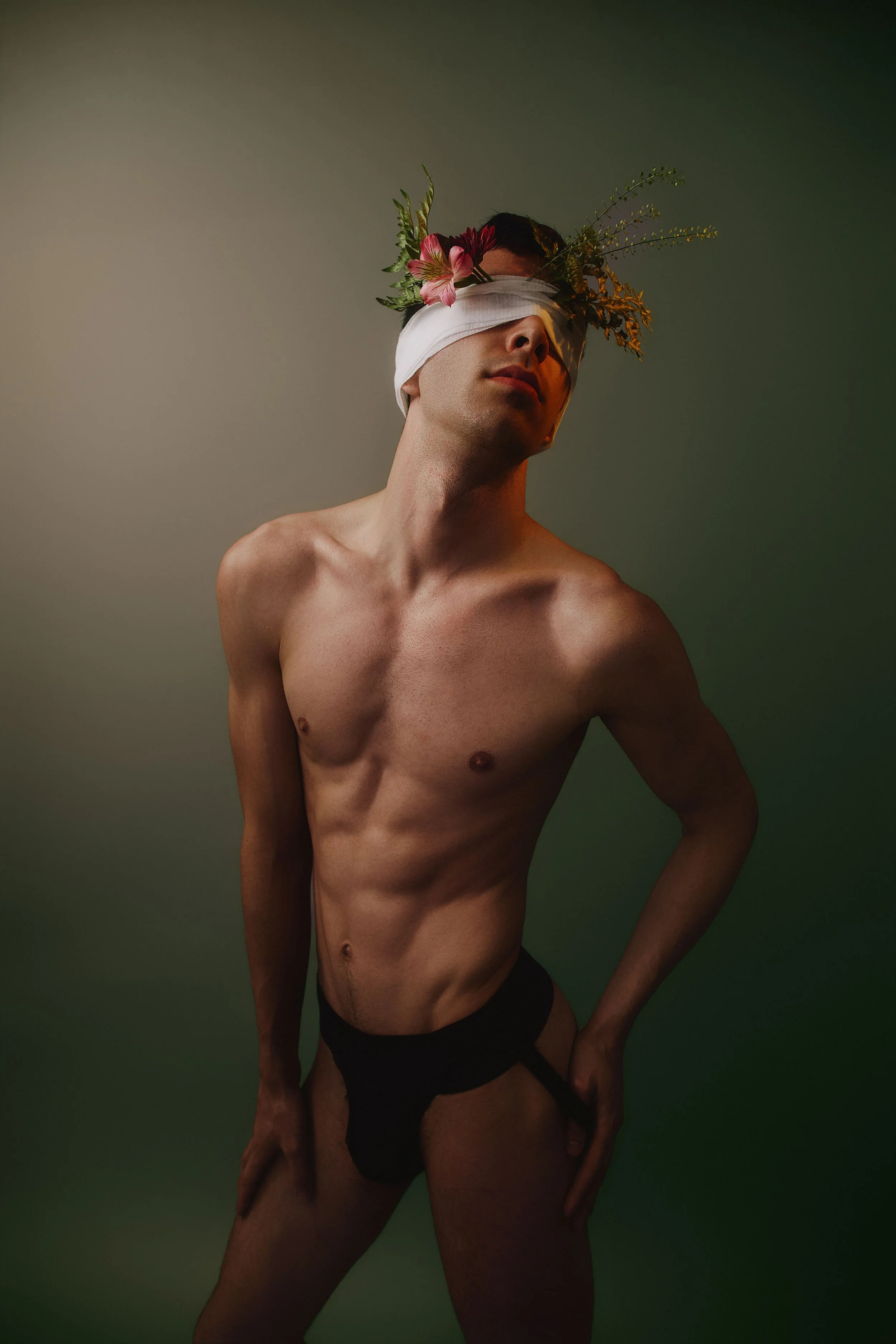 A shirtless man with a blindfold covering his eyes, adorned with pink flowers and green foliage, wearing black underwear, standing against a gradient green background.