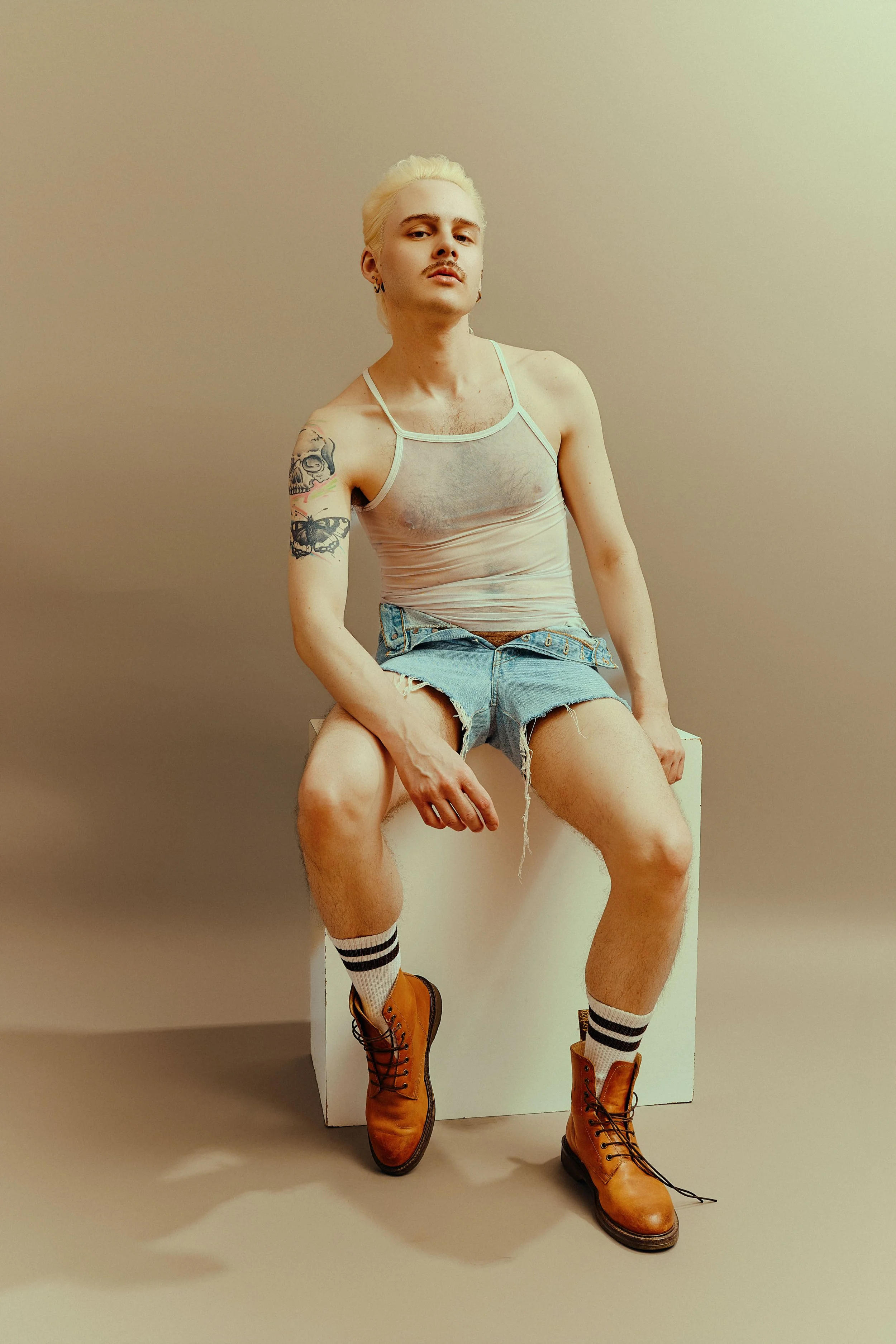 A person with platinum blonde hair, wearing a sheer tank top, denim shorts, striped socks, and brown boots, sitting on a white block against a plain background.