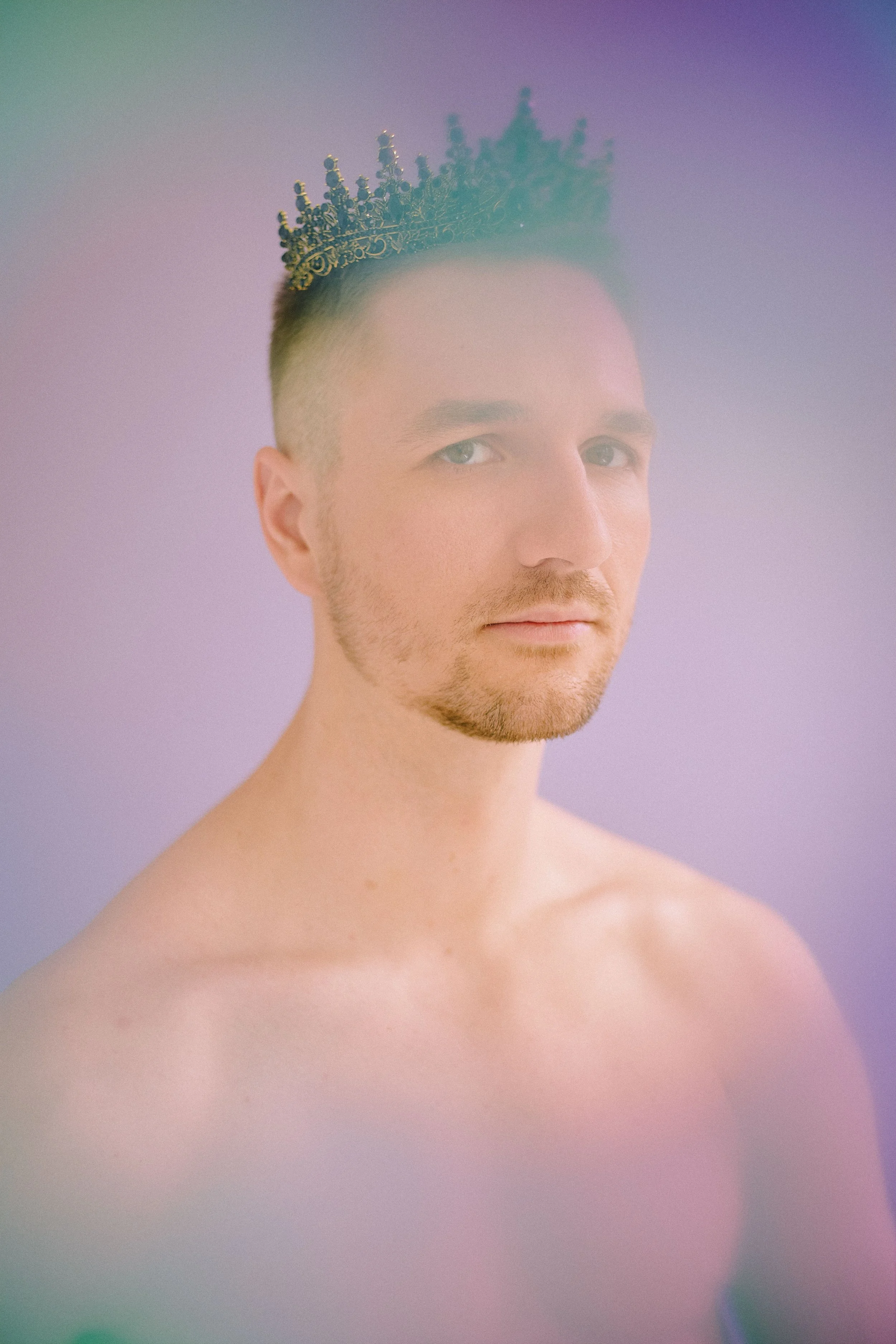 A young man with short, light-colored hair and a beard, wearing a decorative crown, looking at the camera against a softly colored background.