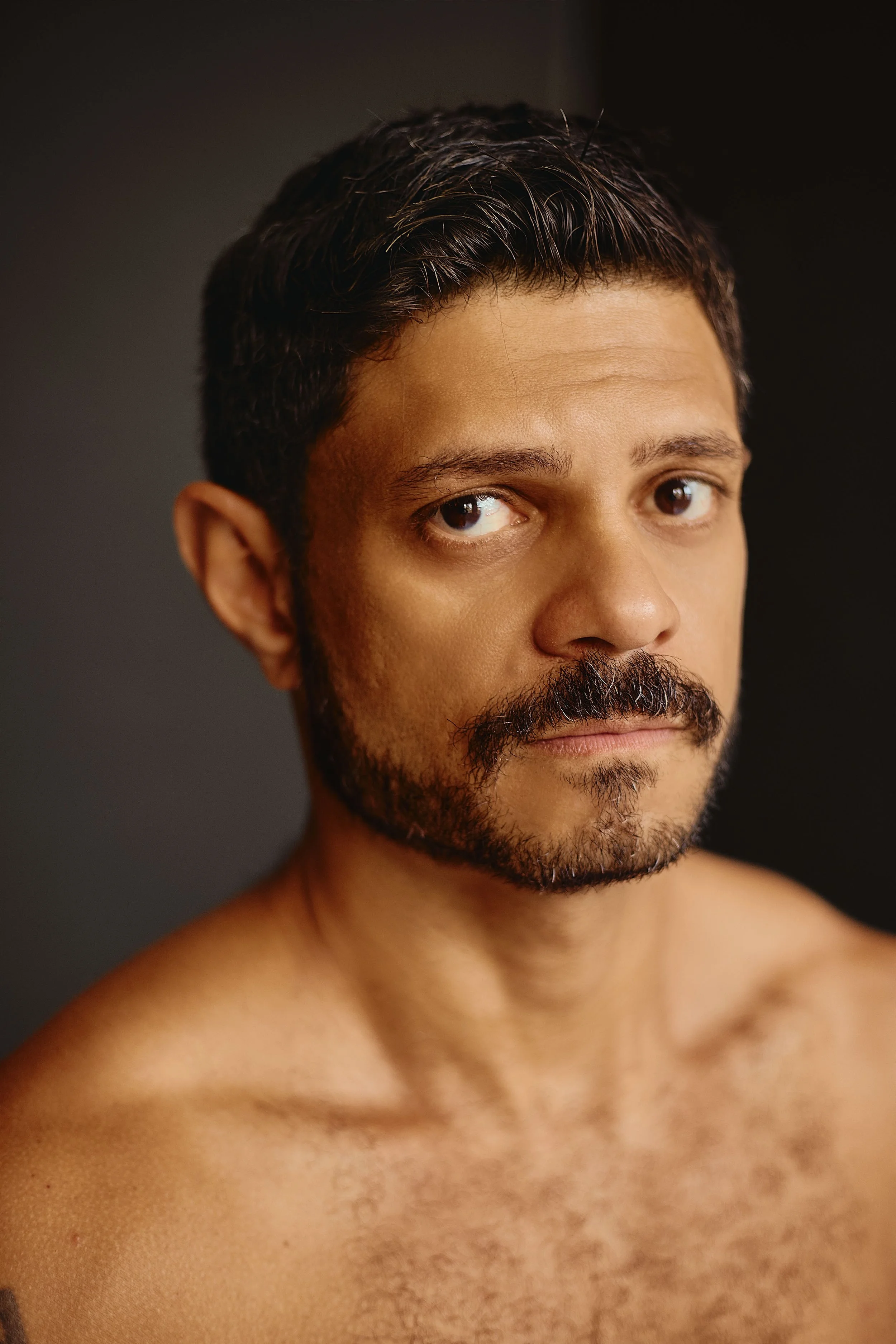 Close-up of a shirtless man with dark hair and a beard, looking to the side against a dark background.