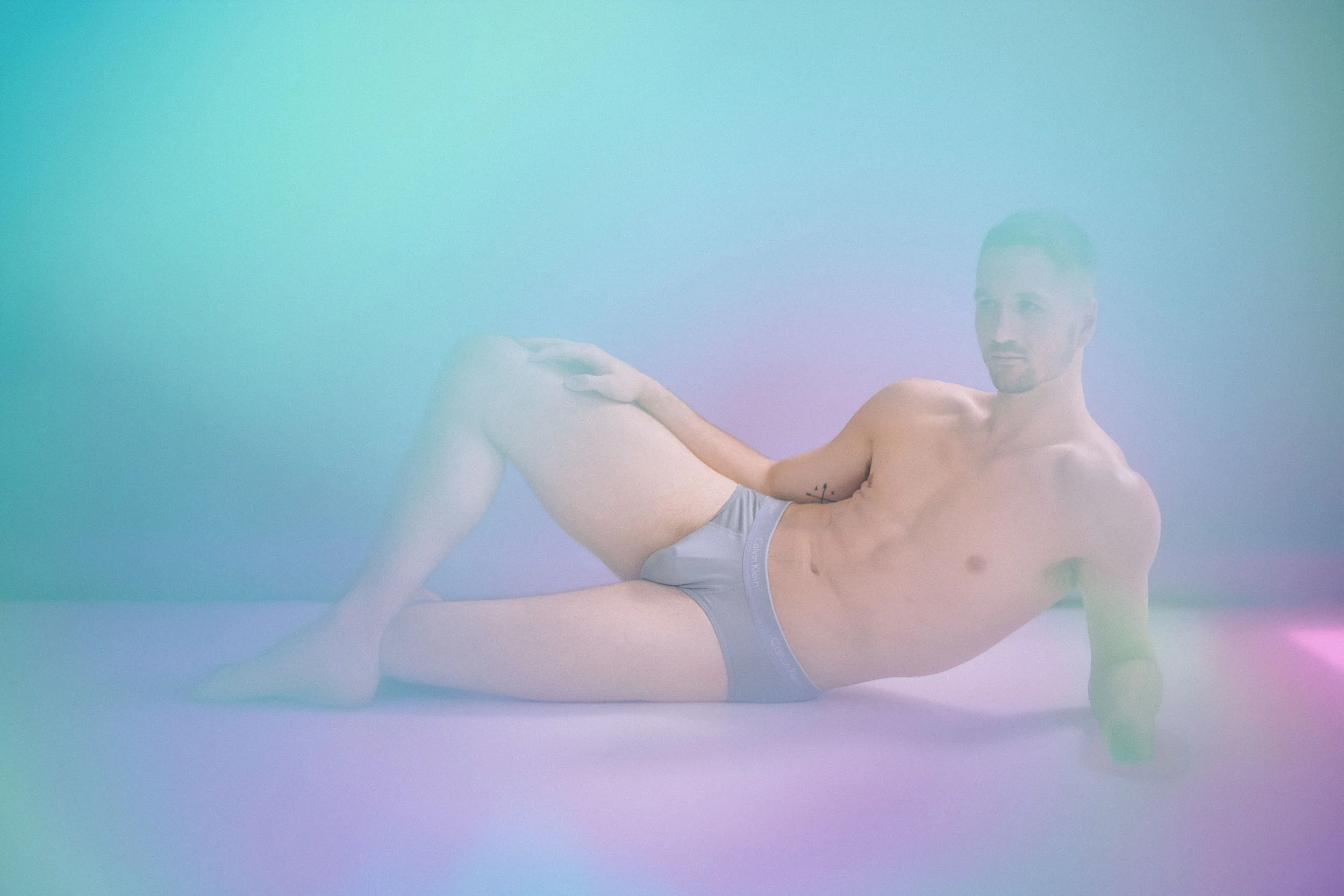 A shirtless man with tattoos on his chest and a tattoo of an anchor on his left arm, wearing gray Calvin Klein underwear, is lying on the ground with his legs raised and bent at the knees, holding his left leg with his right hand. The background is b