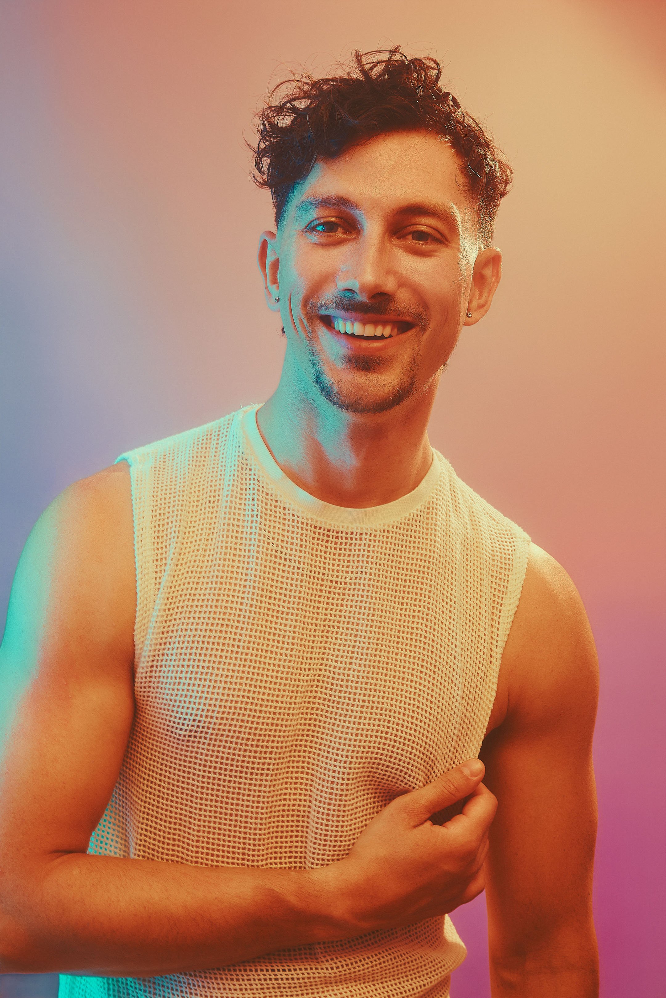 Close-up of a young man smiling, wearing a sleeveless mesh shirt, with colorful lighting in the background.