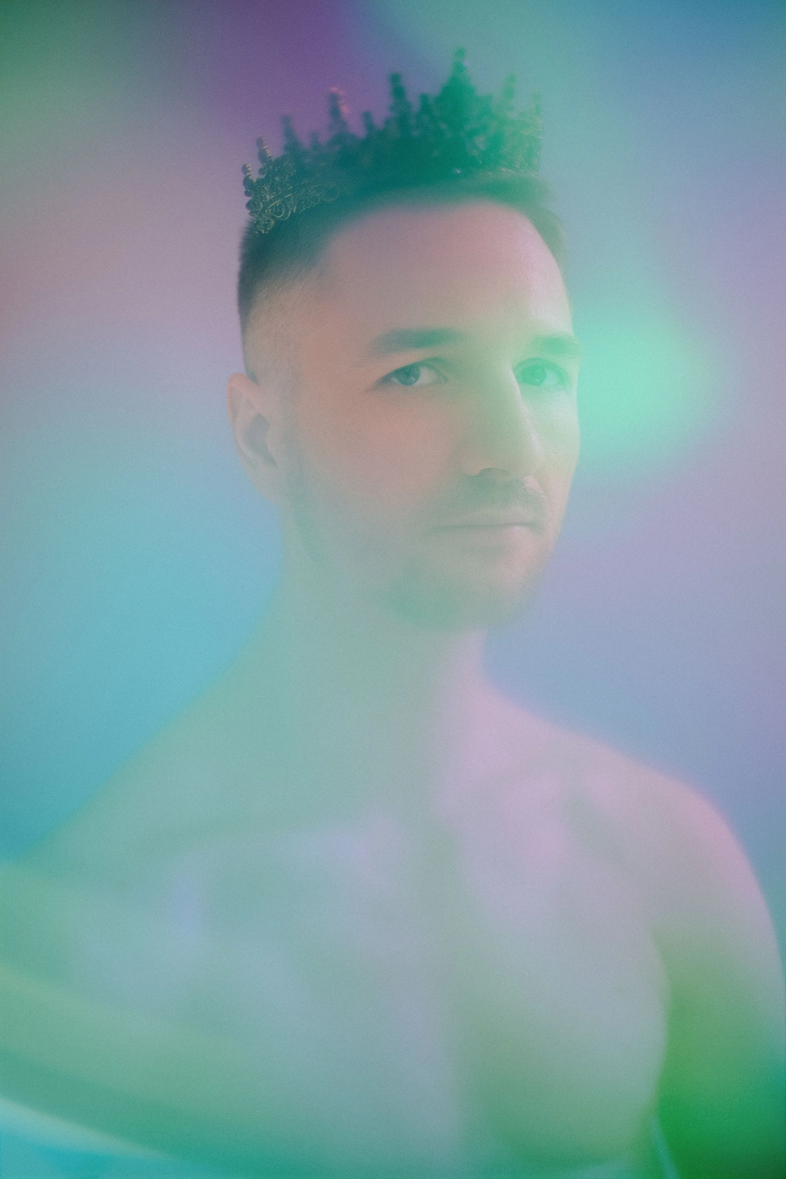 Close-up of a young man with short dark hair, light skin, and blue eyes, wearing a crown, with image overlaid by a colorful, hazy gradient.