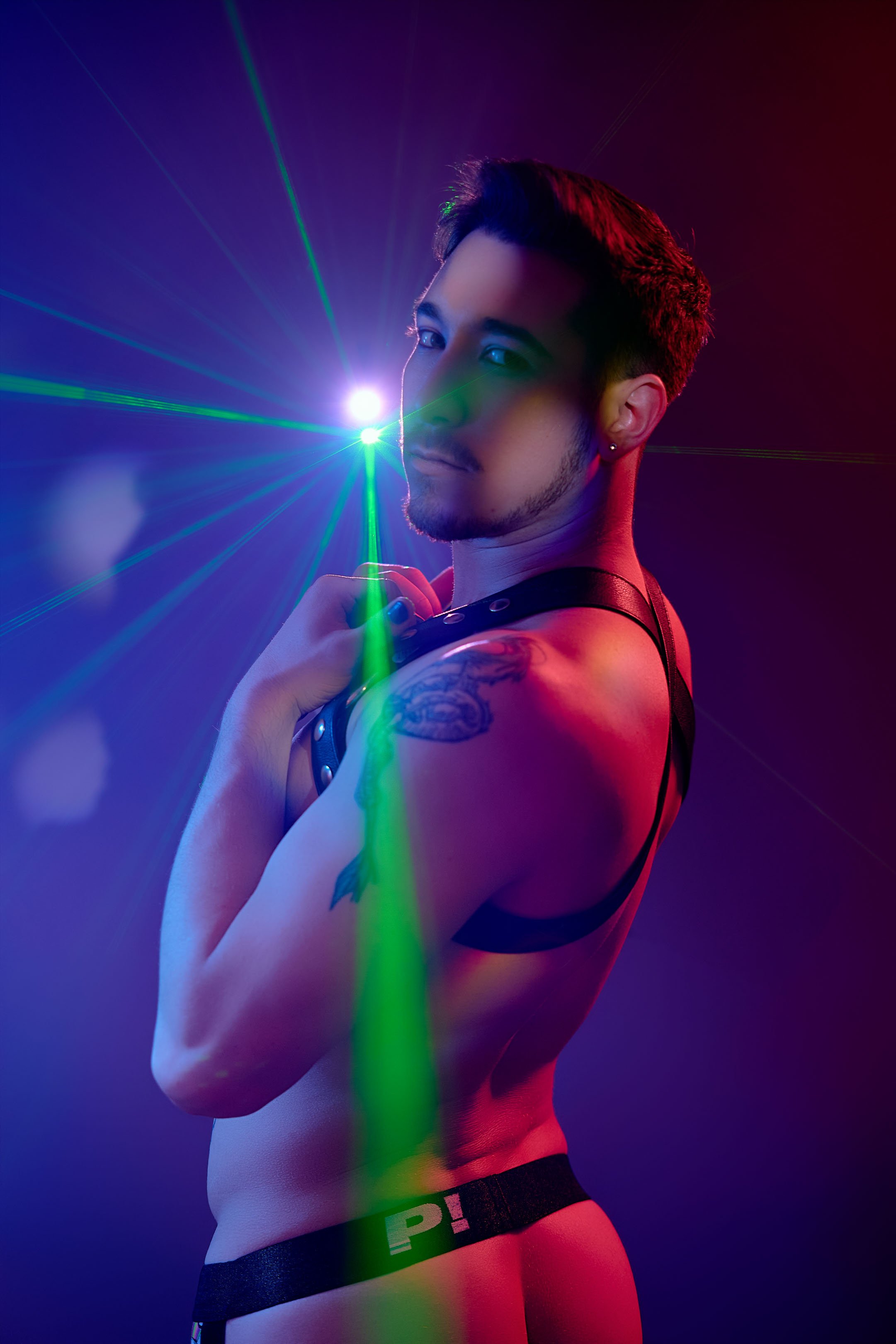 A person with short dark hair, wearing a black harness and black underwear, is standing against a dark background with colorful laser lights, partially illuminated by red and green light.