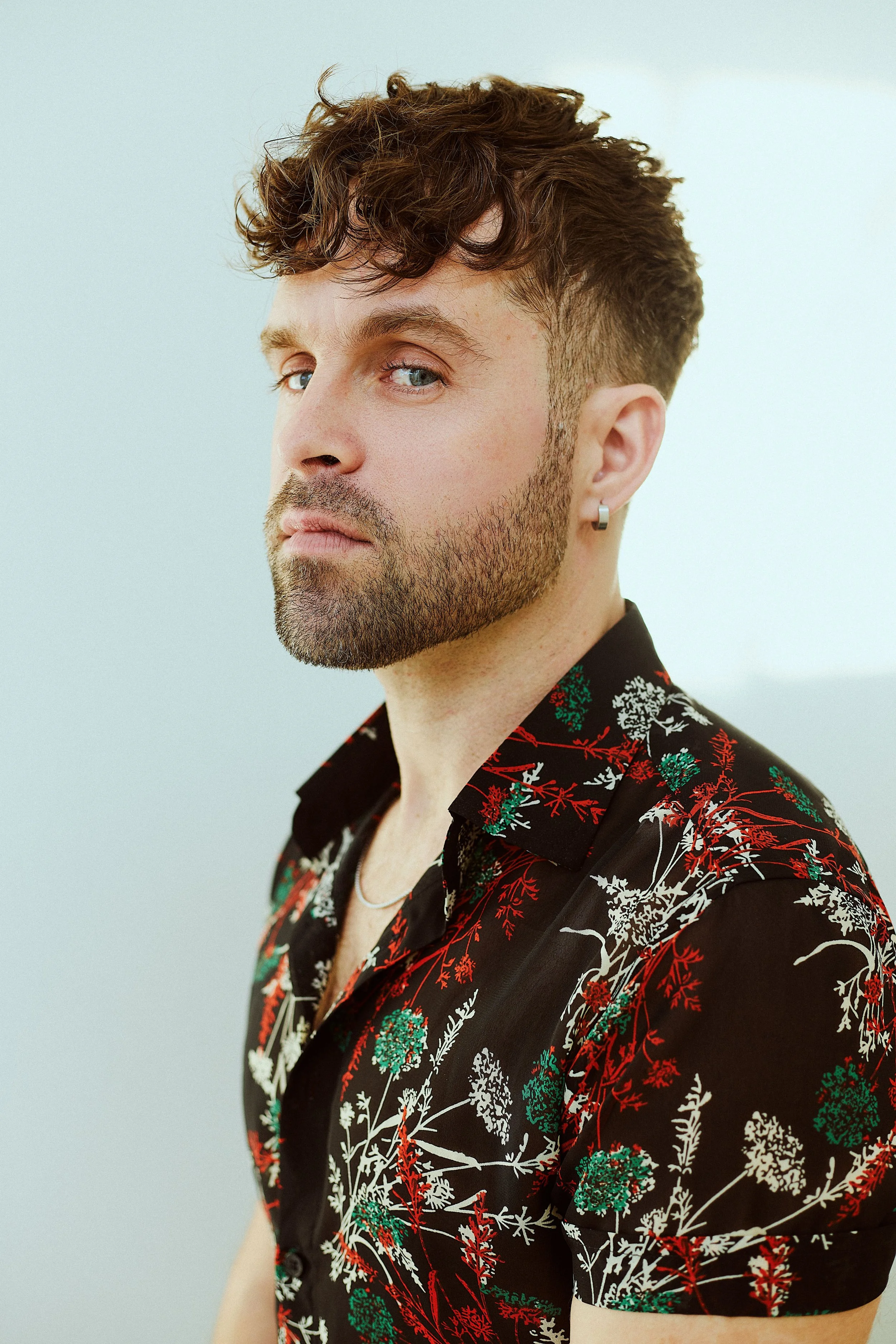 A man with short curly brown hair, blue eyes, and light skin, wearing a black shirt with a colorful floral pattern, standing against a plain light background.
