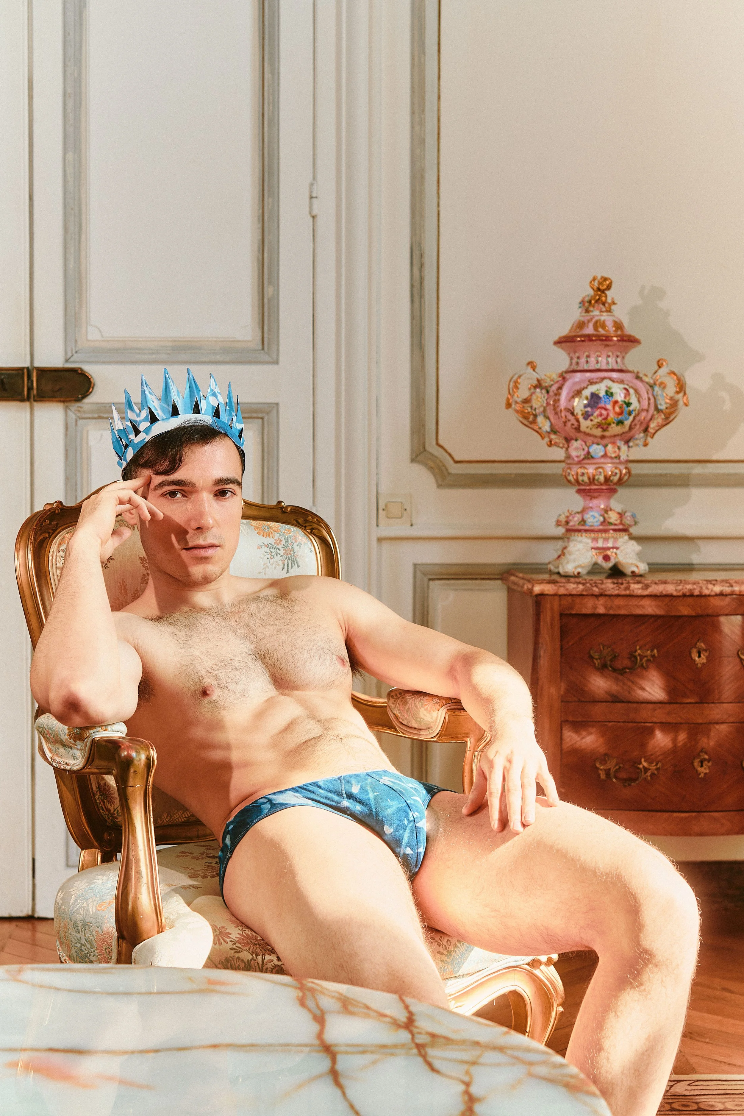 A young man with dark hair, shirtless and wearing blue patterned underwear, poses in a luxurious room with vintage furniture, an ornate pink and floral vase on a side table, and a marble table in the foreground; he wears a blue crown.