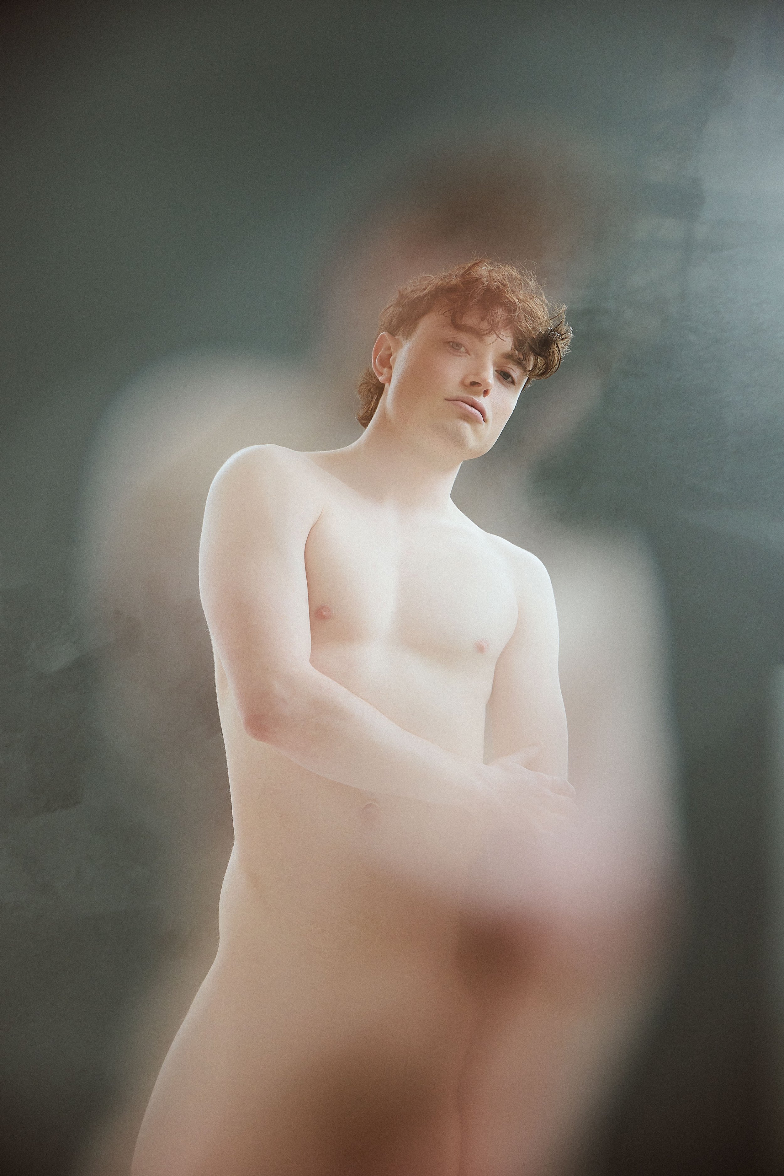 A young person with short, curly brown hair, standing topless with arms crossed in front of their torso, against a blurred, abstract background.