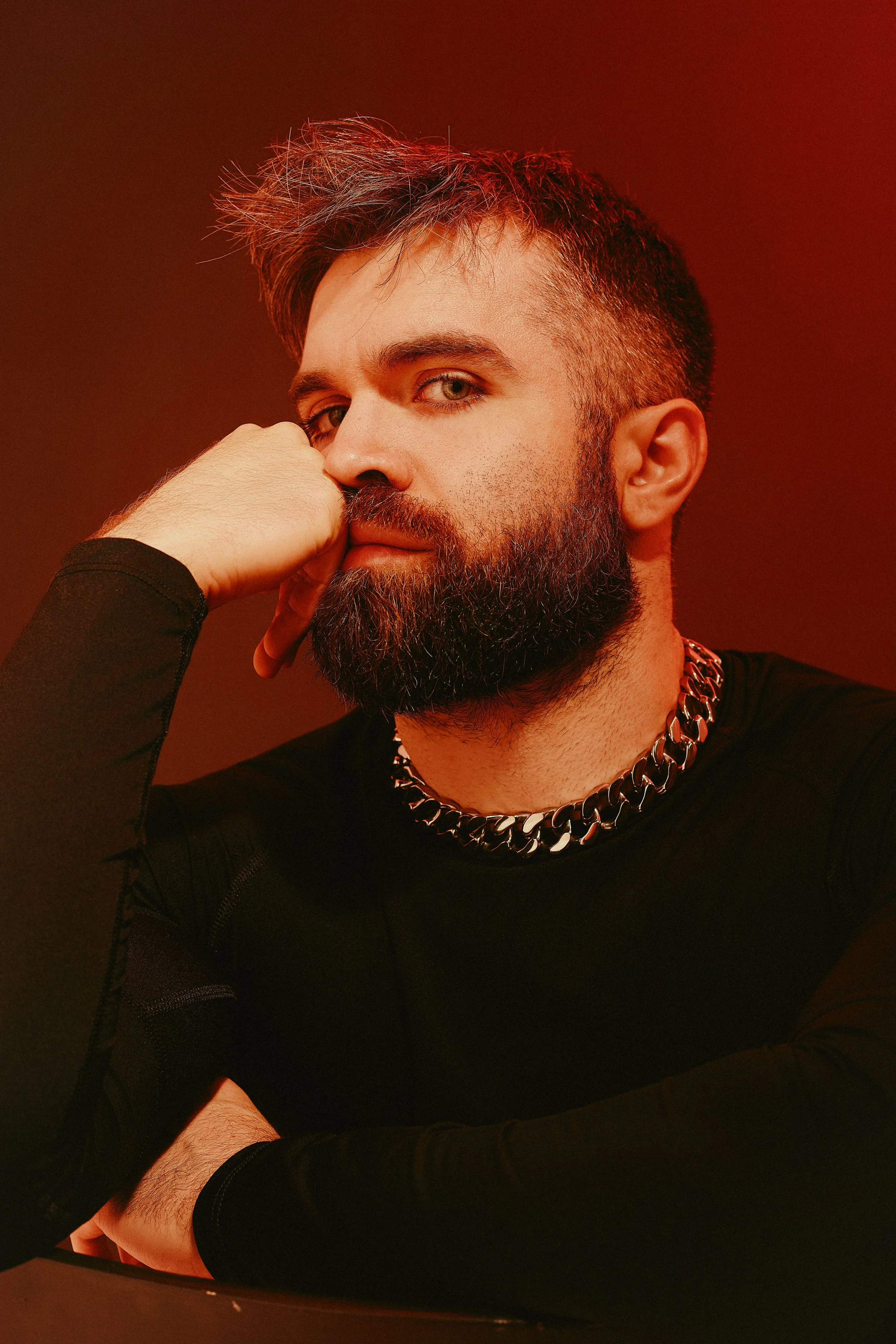 A man with a full beard and styled hair wearing a black shirt and a thick chain necklace, resting his face on his hand, in a red-lit setting.