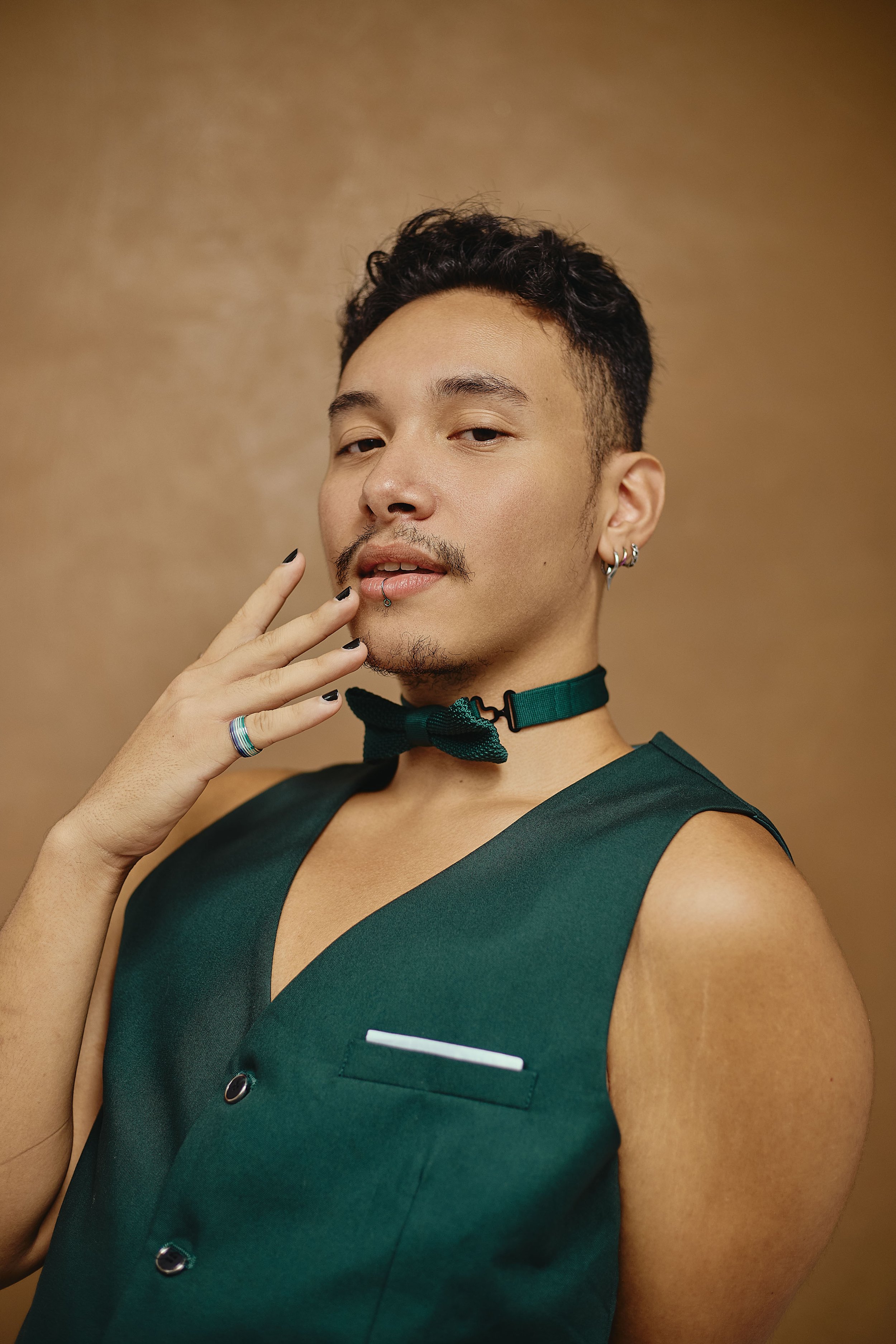A person with short dark hair, wearing a green vest and a bow tie, posing against a plain brown background. They are touching their face with one hand, which has rings and a lip piercing, and are wearing earrings.
