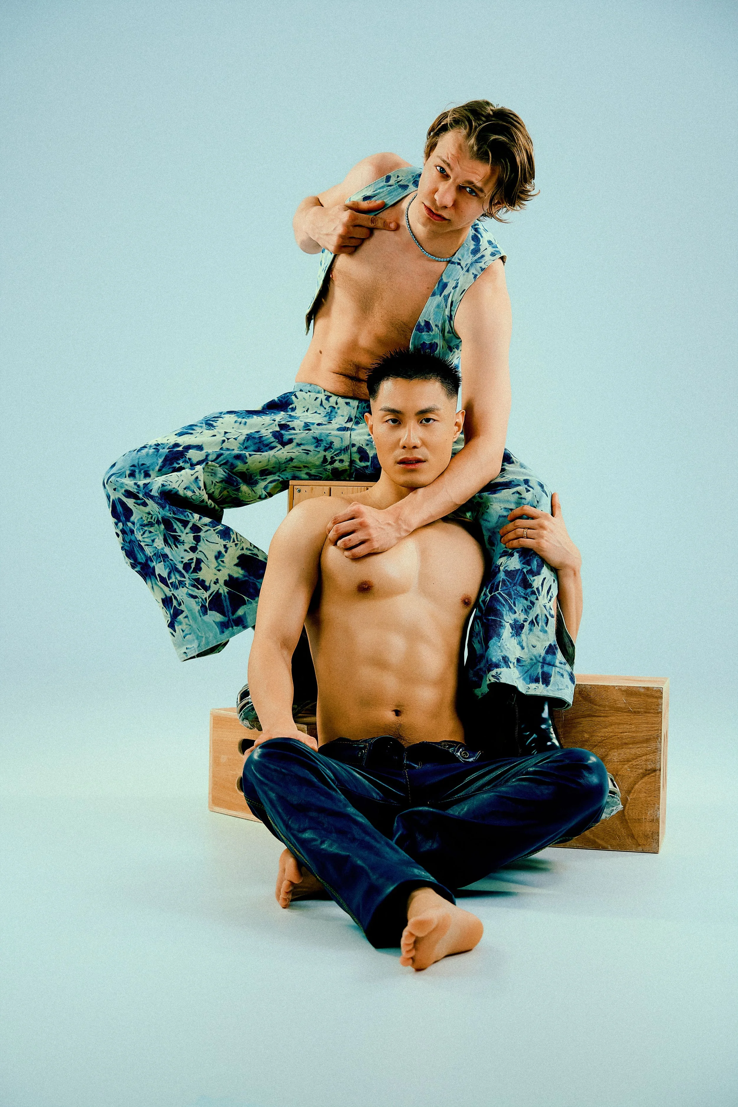 Three young men with light skin and dark hair, posing together in a studio, with one sitting on the ground and the other two standing behind him, wearing casual clothing and accessories.