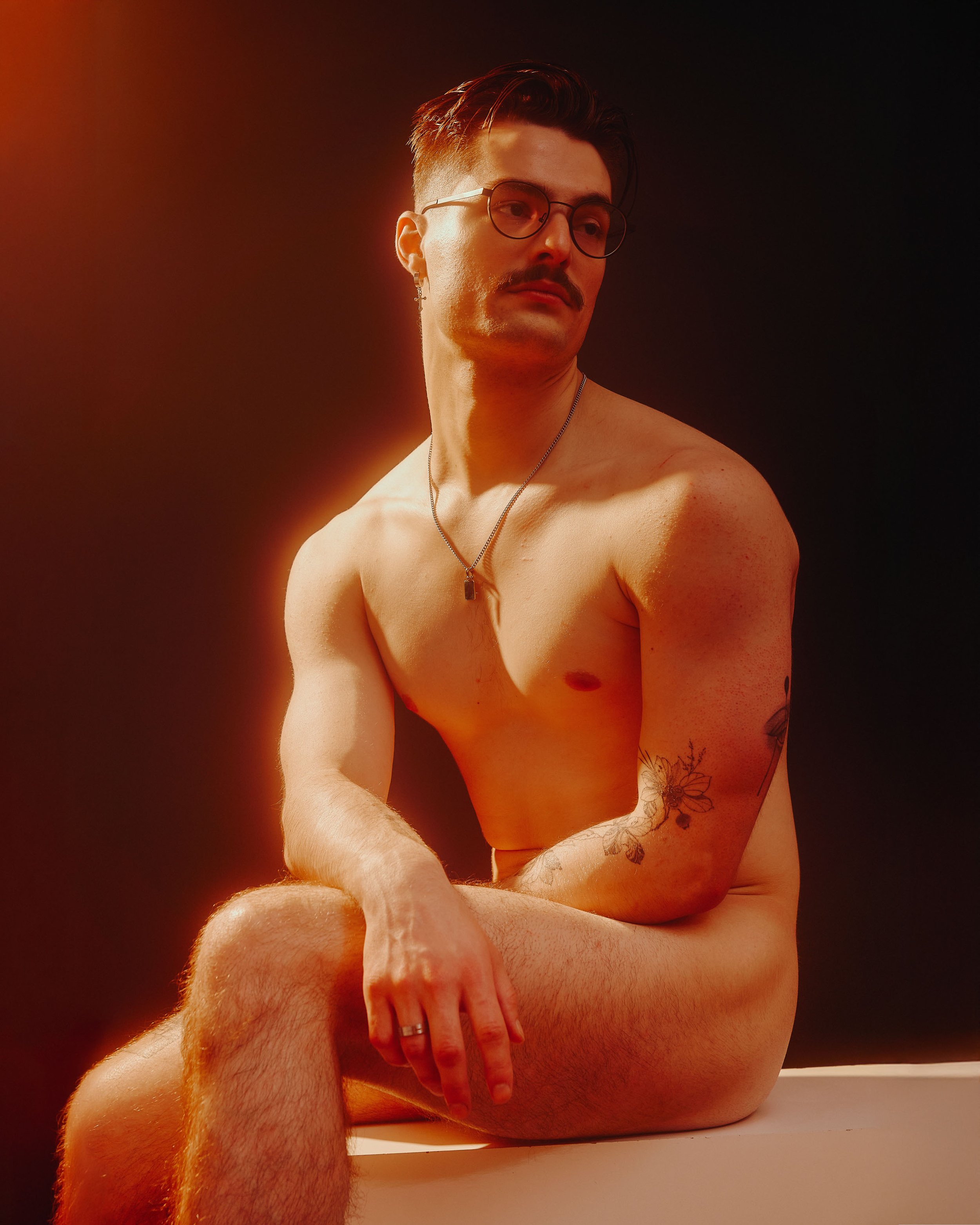 A shirtless man with glasses and a mustache sitting on a white surface against a dark background with warm light highlighting parts of his face and body, showing tattoos on his left arm.