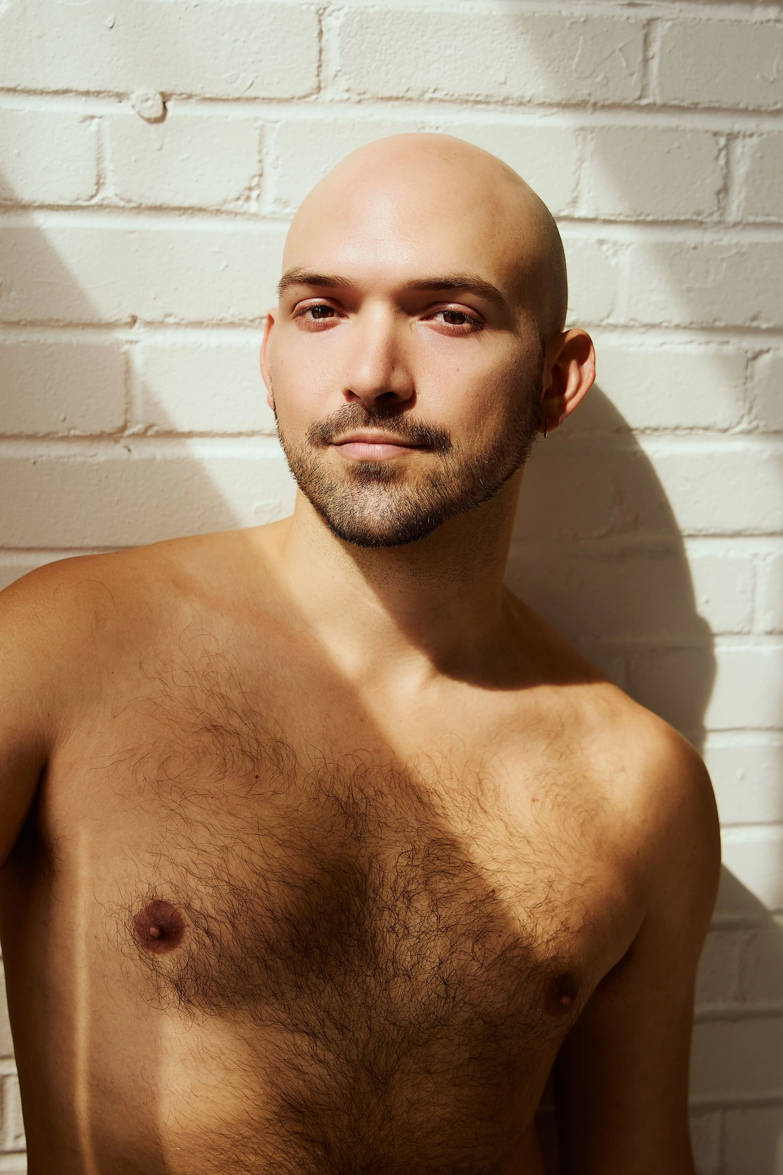 A shirtless man with a beard and short hair, leaning against a white brick wall, with sunlight casting shadows on his face and chest.