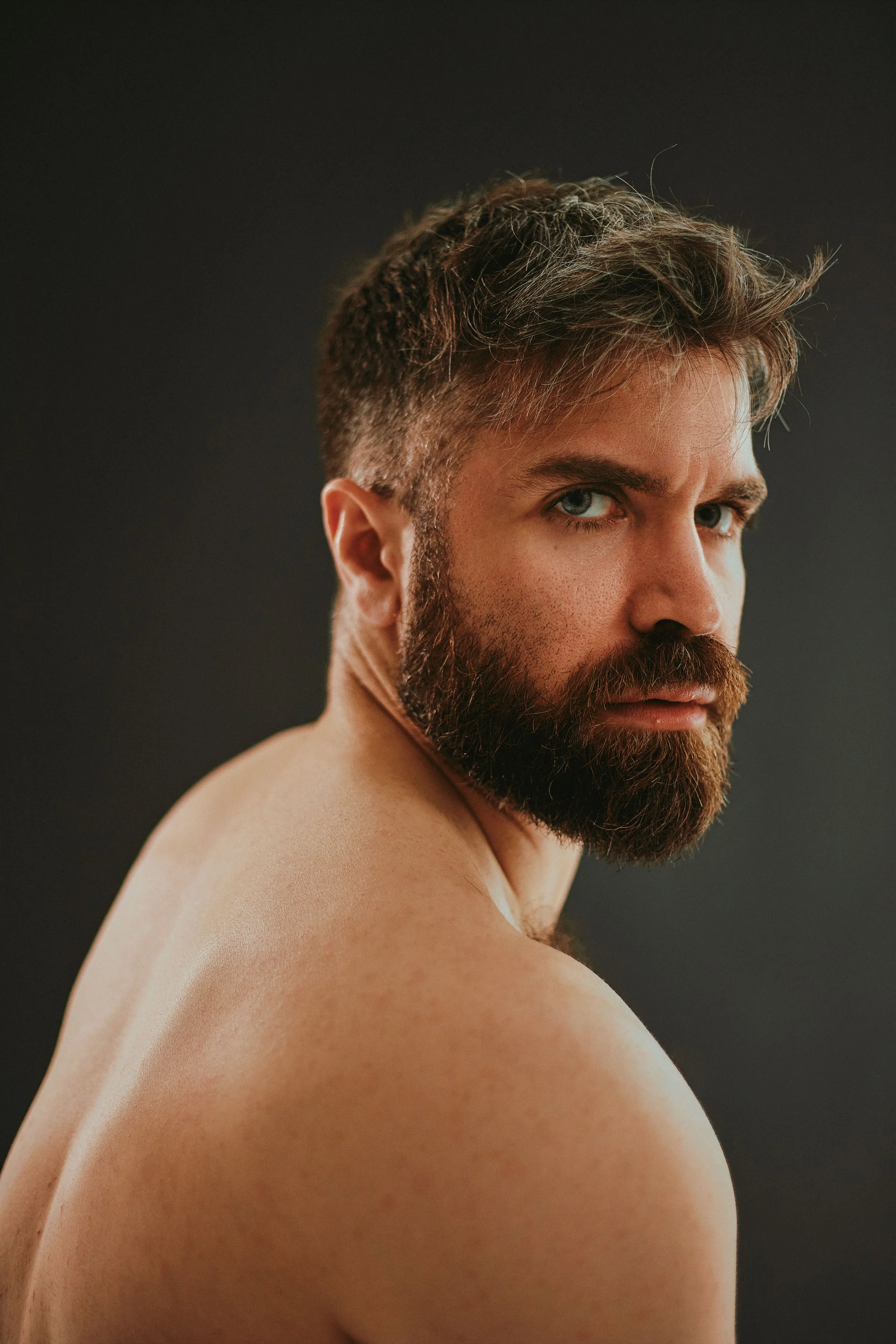 Close-up of a shirtless man with a thick beard and styled hair, looking over his shoulder against a dark background.