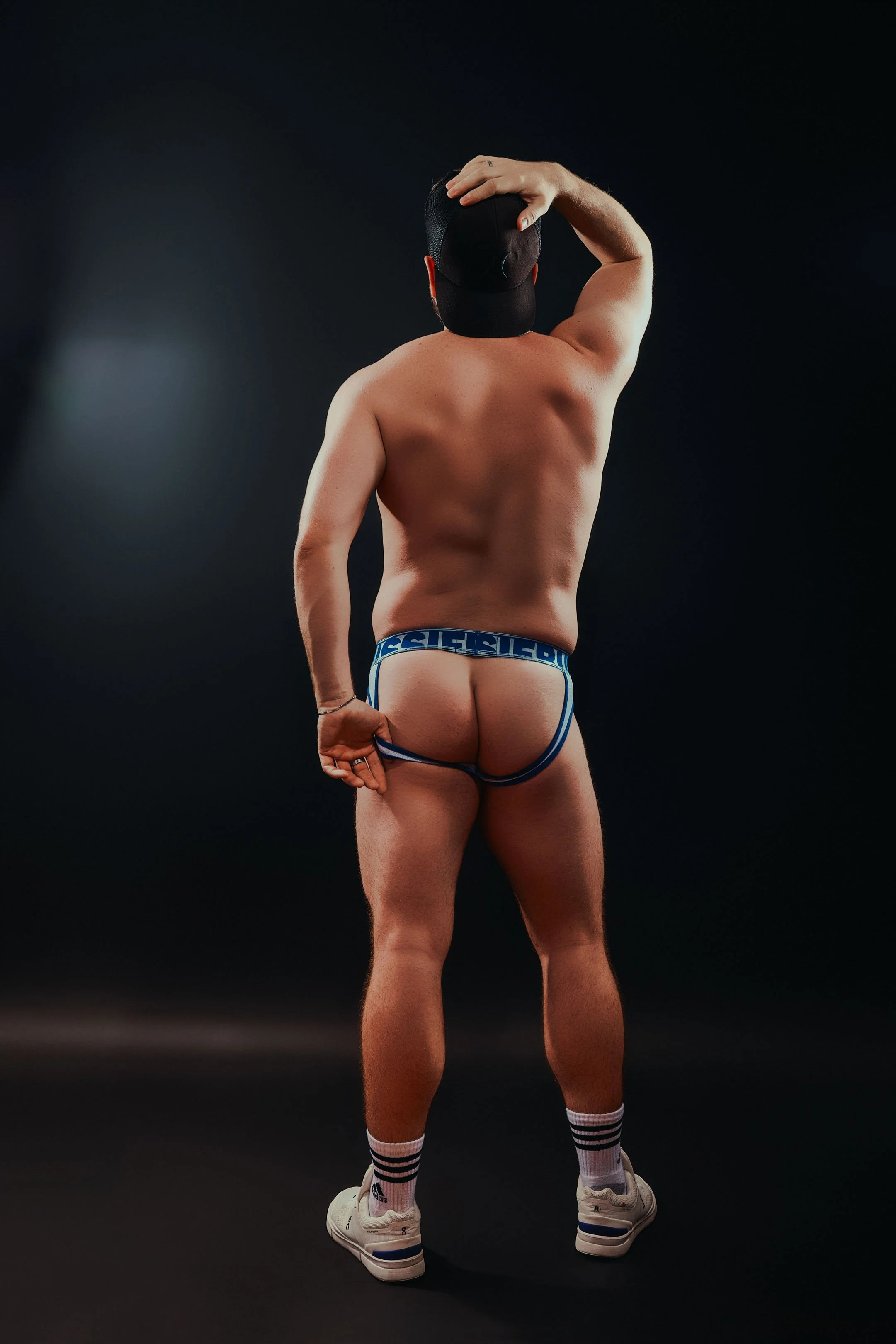 A shirtless man wearing a black cap, striped socks, white sneakers, and blue briefs with the word 'SIE' visible, standing against a dark background with his back to the camera, touching his head with one hand.