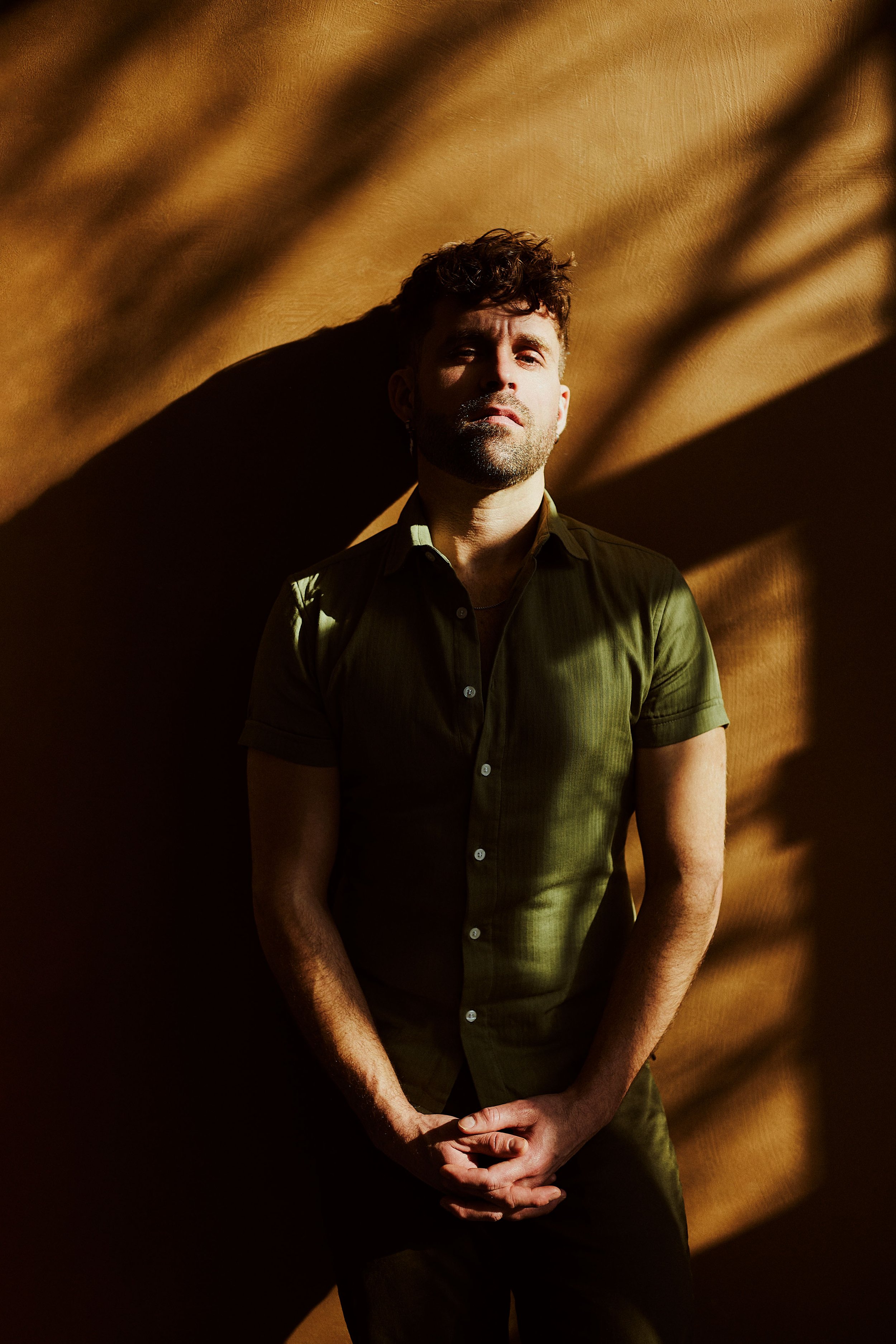 A man with curly hair and a beard stands against a textured, warm-toned background, partially in shadow, with his hands clasped in front of him.