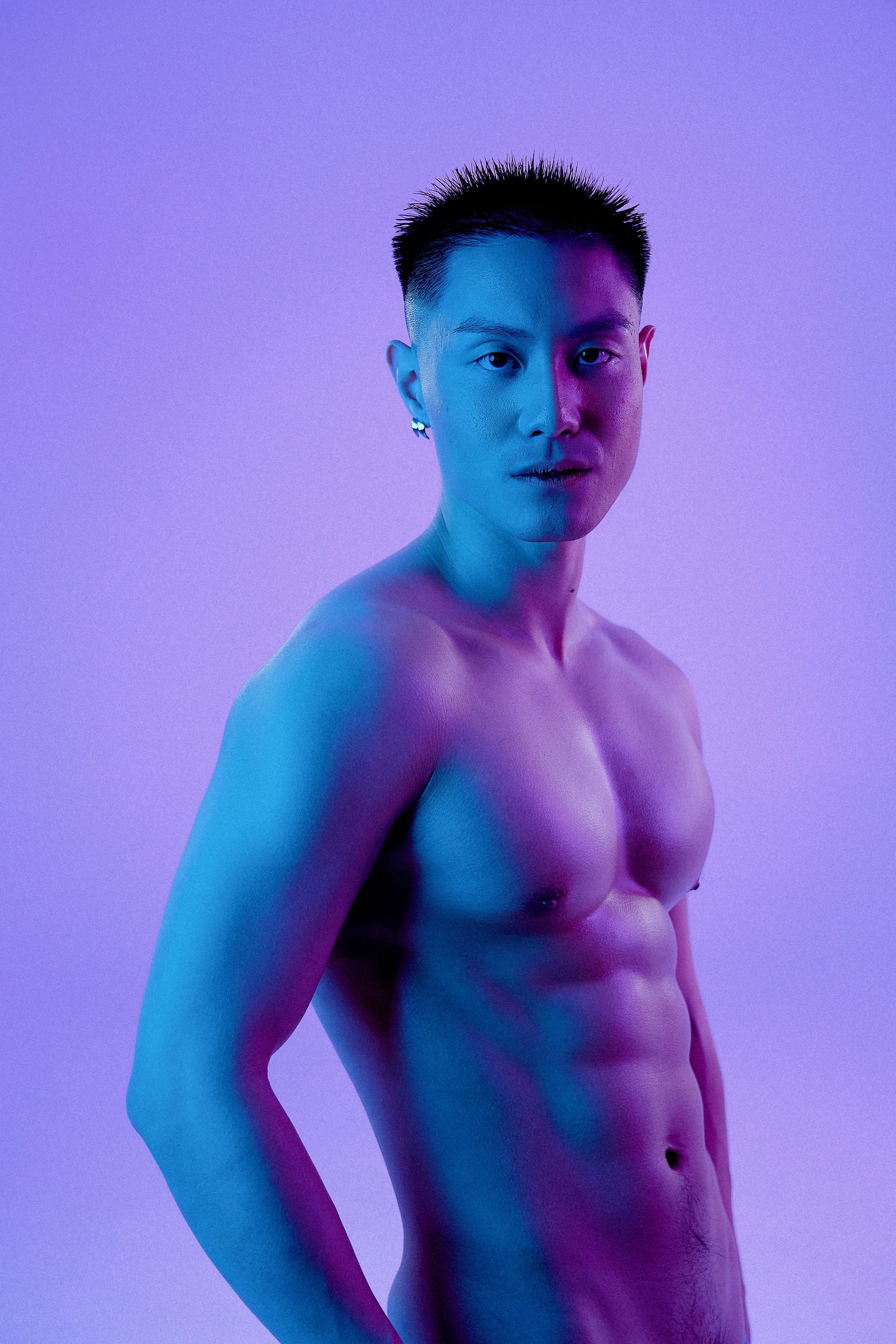 A shirtless person with short black hair and earrings, illuminated with blue and purple lighting, standing against a purple background.