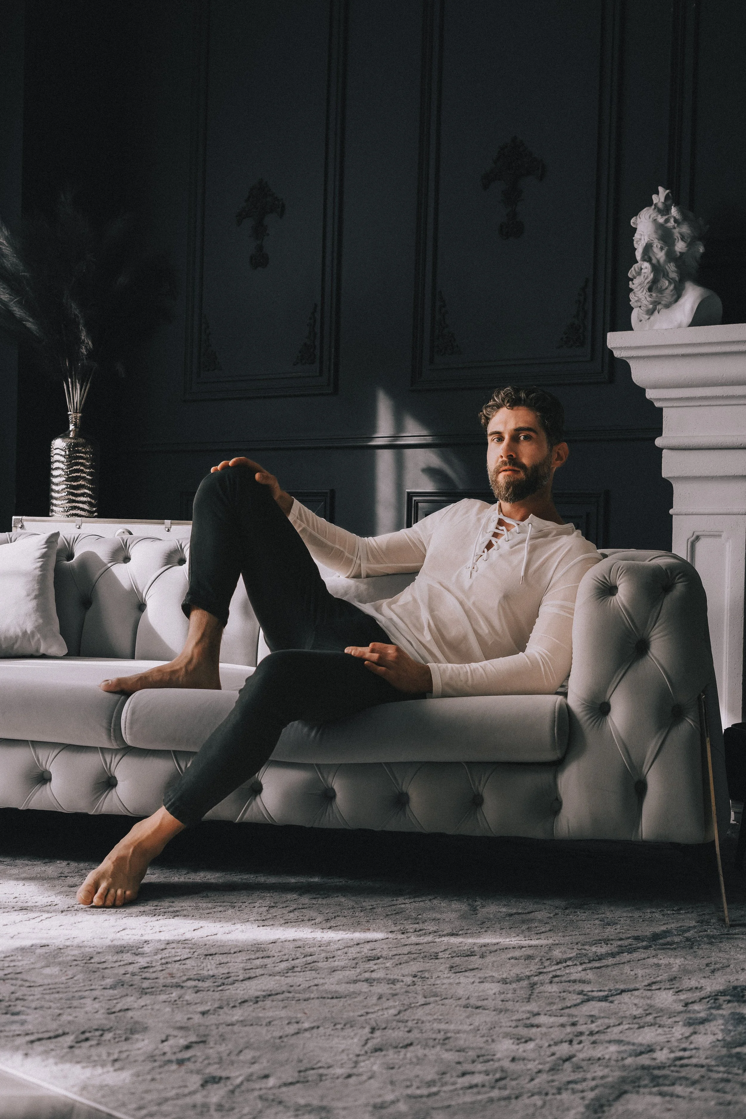 A man with a beard and curly hair is reclining on a white tufted sofa in a dark, elegant room with black-paneled walls and a sculpted bust of a lion on the fireplace mantel. He is wearing a light-colored long-sleeve shirt and dark pants, with his lef