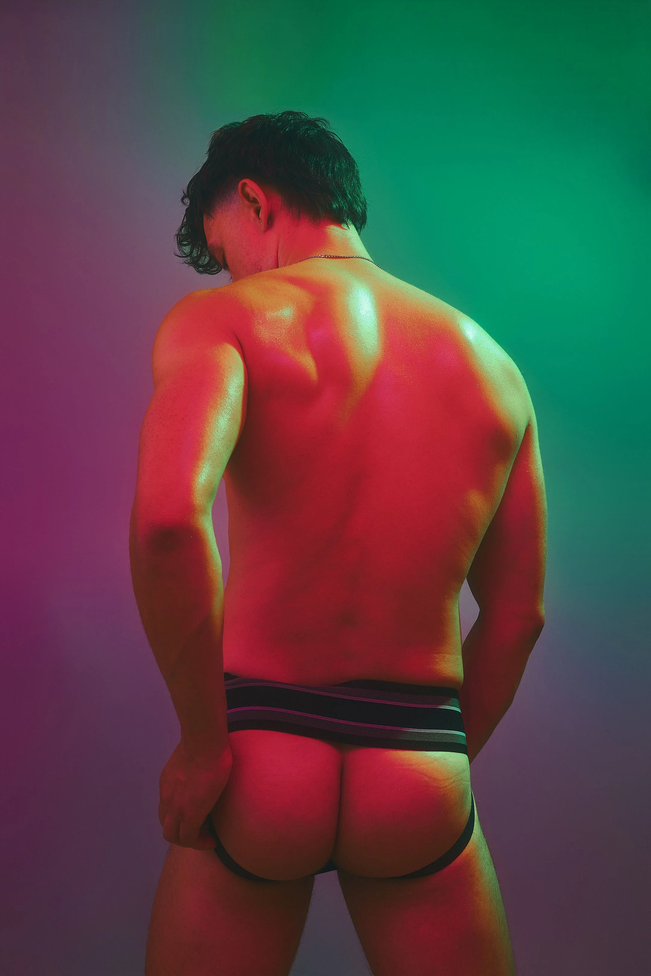 A person with short dark hair, showing their back and wearing striped boxer briefs, illuminated with multicolored neon lighting.