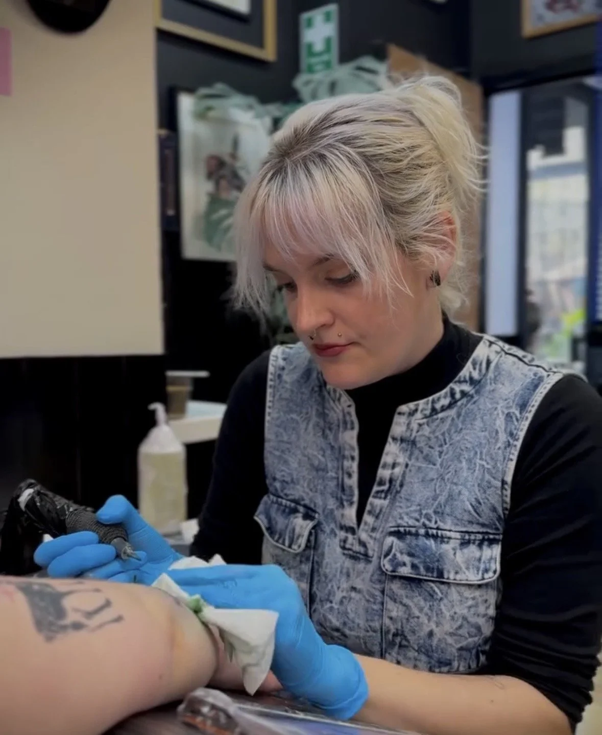 A woman receives a tattoo on her arm from a tattoo artist wearing blue gloves in a tattoo shop.