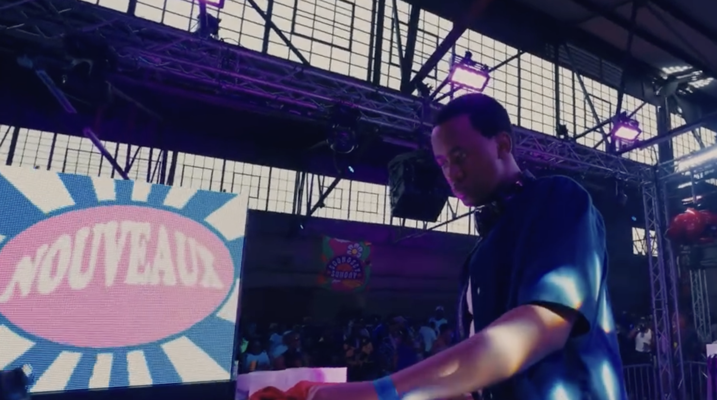A DJ performing at an indoor event with purple lighting, standing behind a DJ booth that has a colorful logo, under a metal framework with large windows in the background.