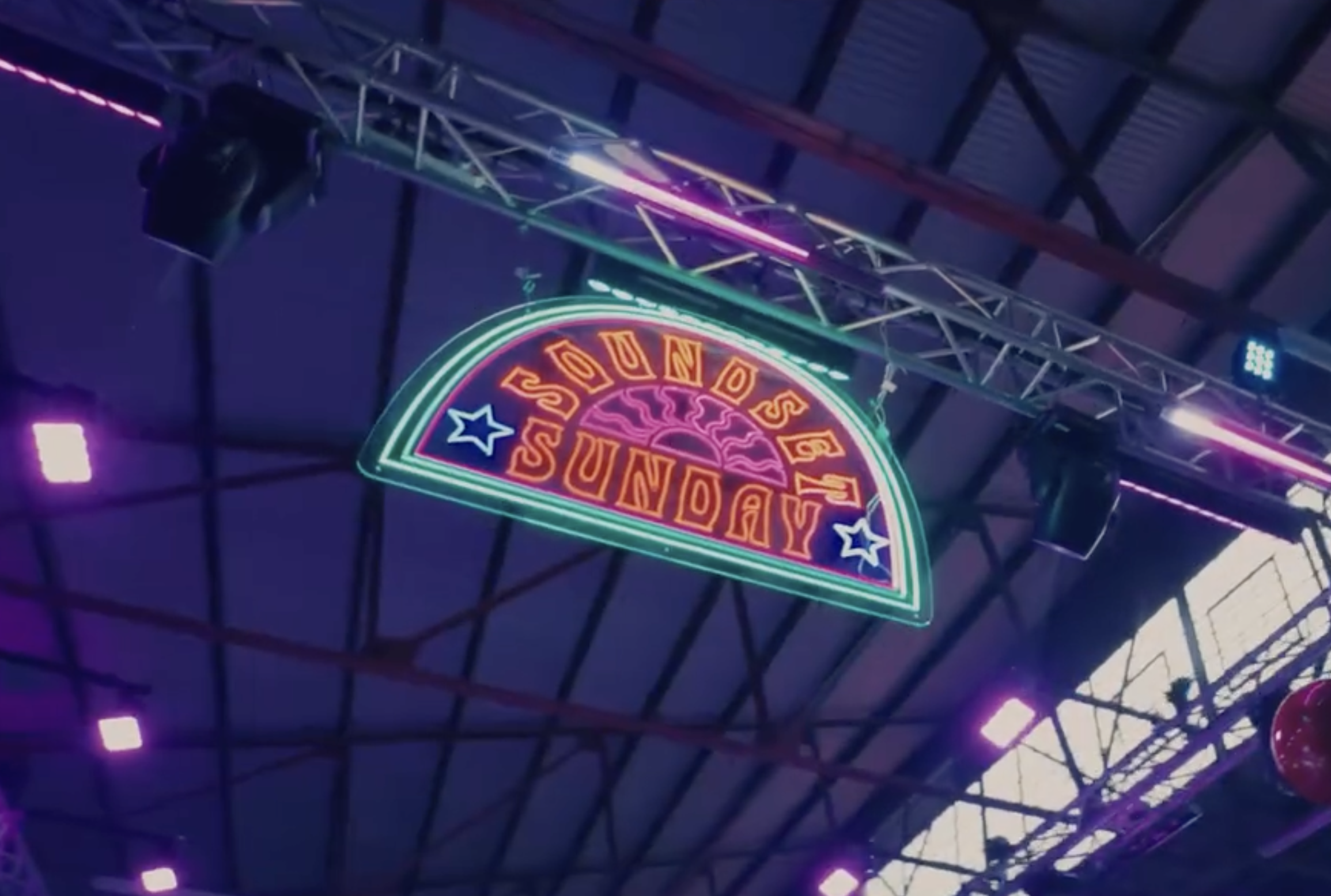 Colorful neon sign with the words 'SOUND SUNDAY' hanging inside a large industrial-style building with a high ceiling and metal framework.