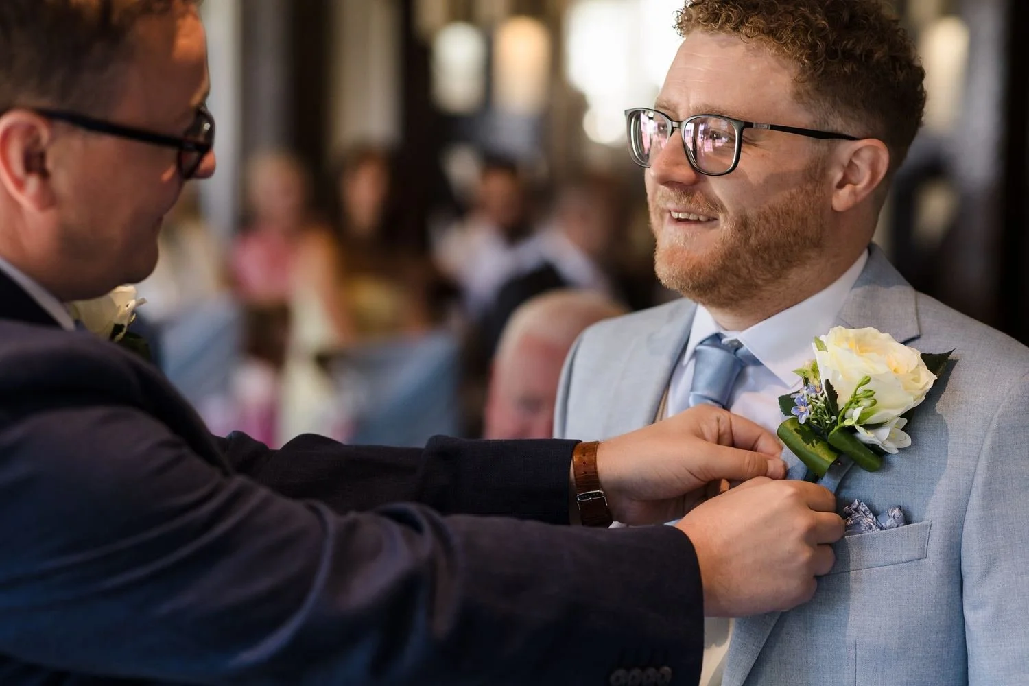 Josh smiling as his white rose buttonhole is pinned to his pale blue jacket before the ceremony