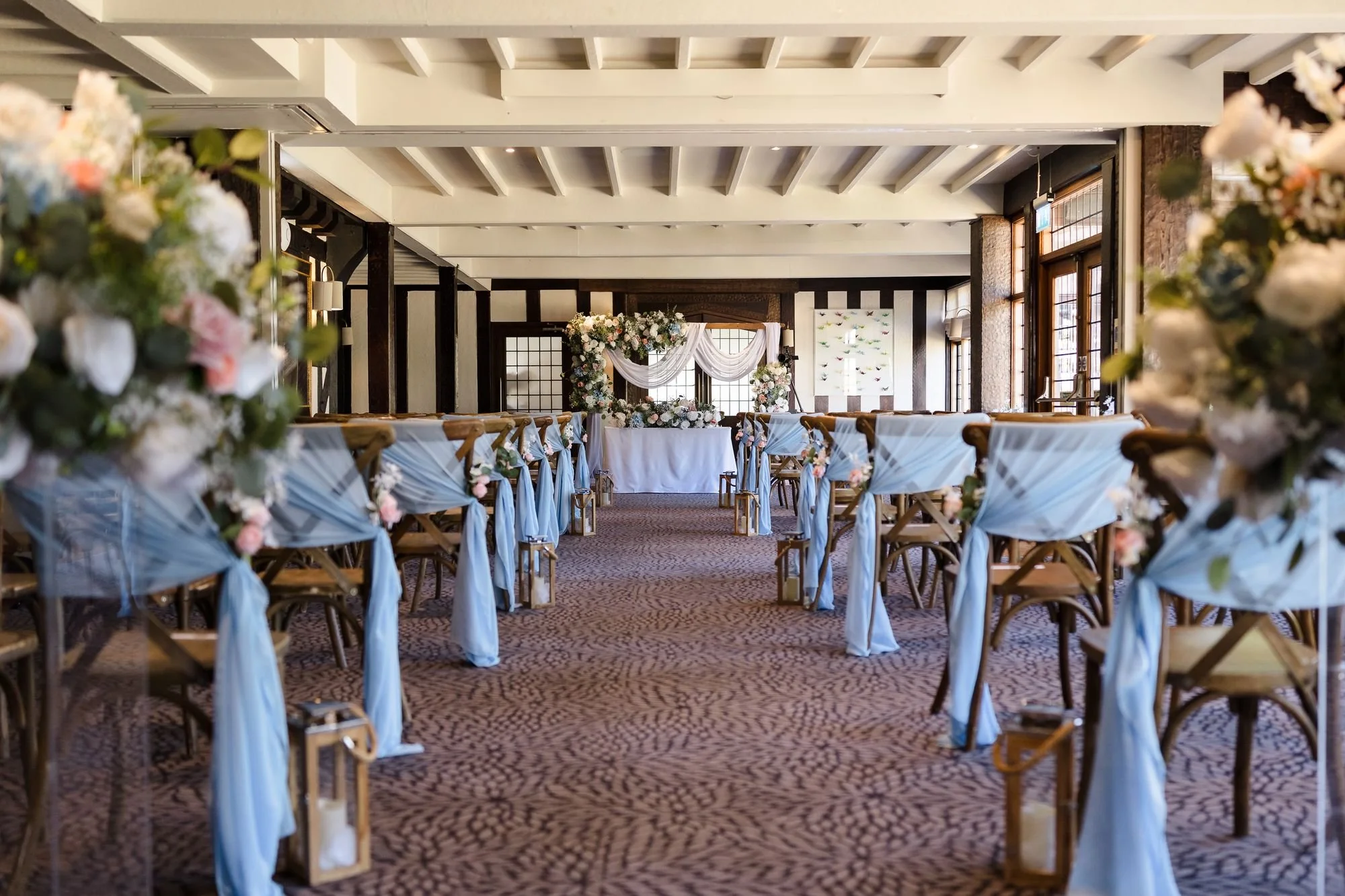 Drawing Room ceremony aisle at Hogarths Stone Manor with sky-blue chair drapes, lanterns and a floral arch