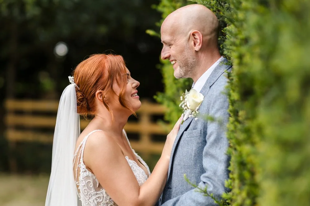 Bride and groom laughing together outdoors at Shustoke Barn, bride in lace wedding gown with veil and red hair up, groom in light grey suit with white rose buttonhole, framed by lush green foliage