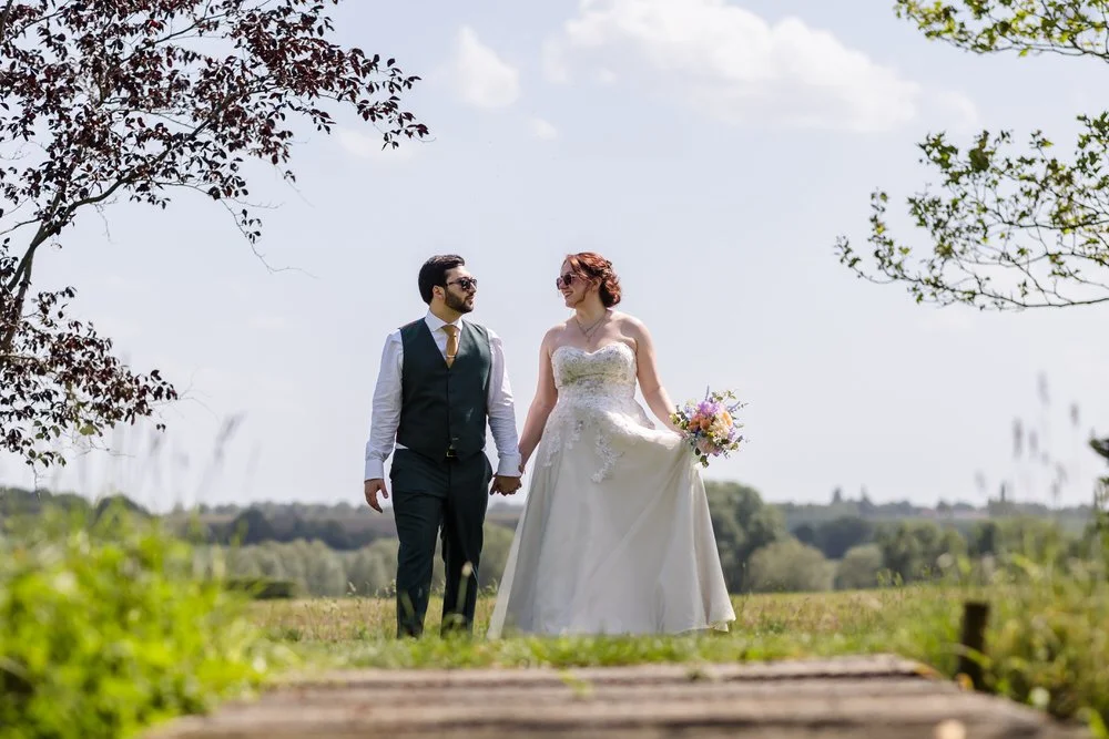 Bride and groom walking hand in hand along a path at Huntsmill Farm, fields beyond.