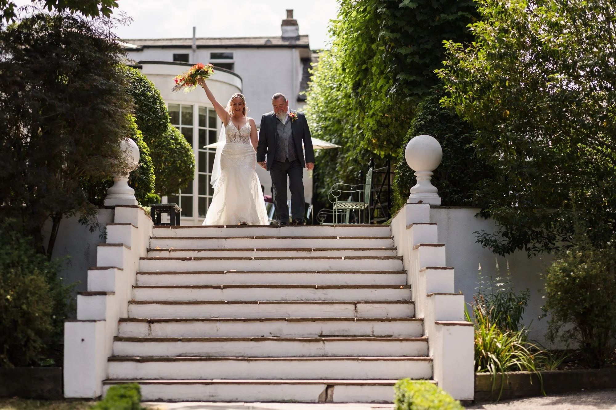 Bride raising her bouquet at the top of the garden steps as she arrives with her dad for her Warwick House ceremony