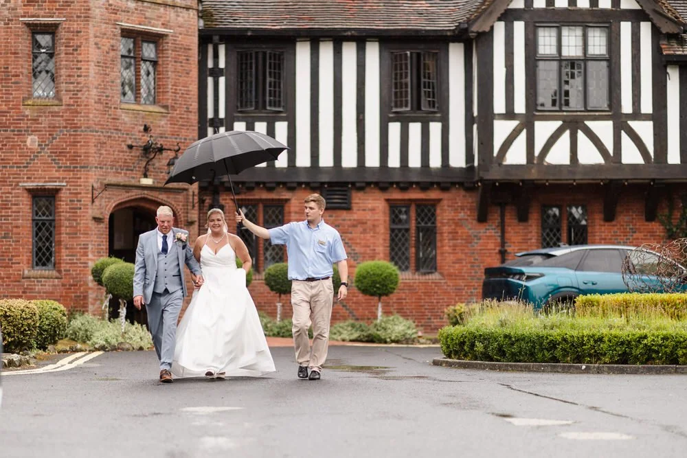 Jess and Brian walking in rain outside Hogarths Stone Manor with umbrella, documentary wedding photography