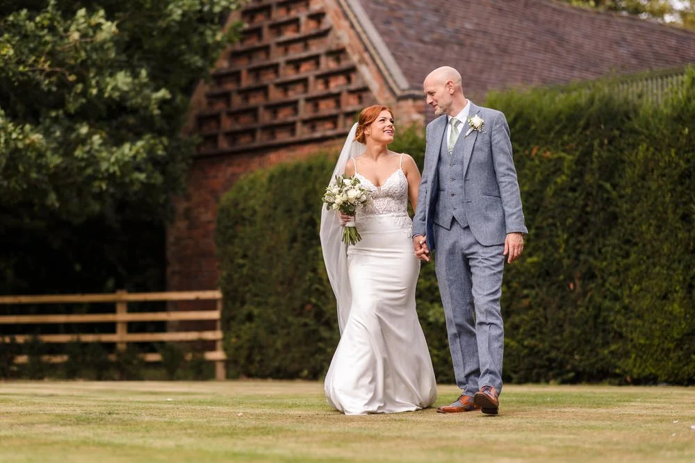 Bride and groom walking hand in hand across the lawns at Shustoke Barn, bride in fitted lace wedding gown carrying a white rose bouquet, groom in light grey three-piece suit with sage green tie, historic brick barn building in the background