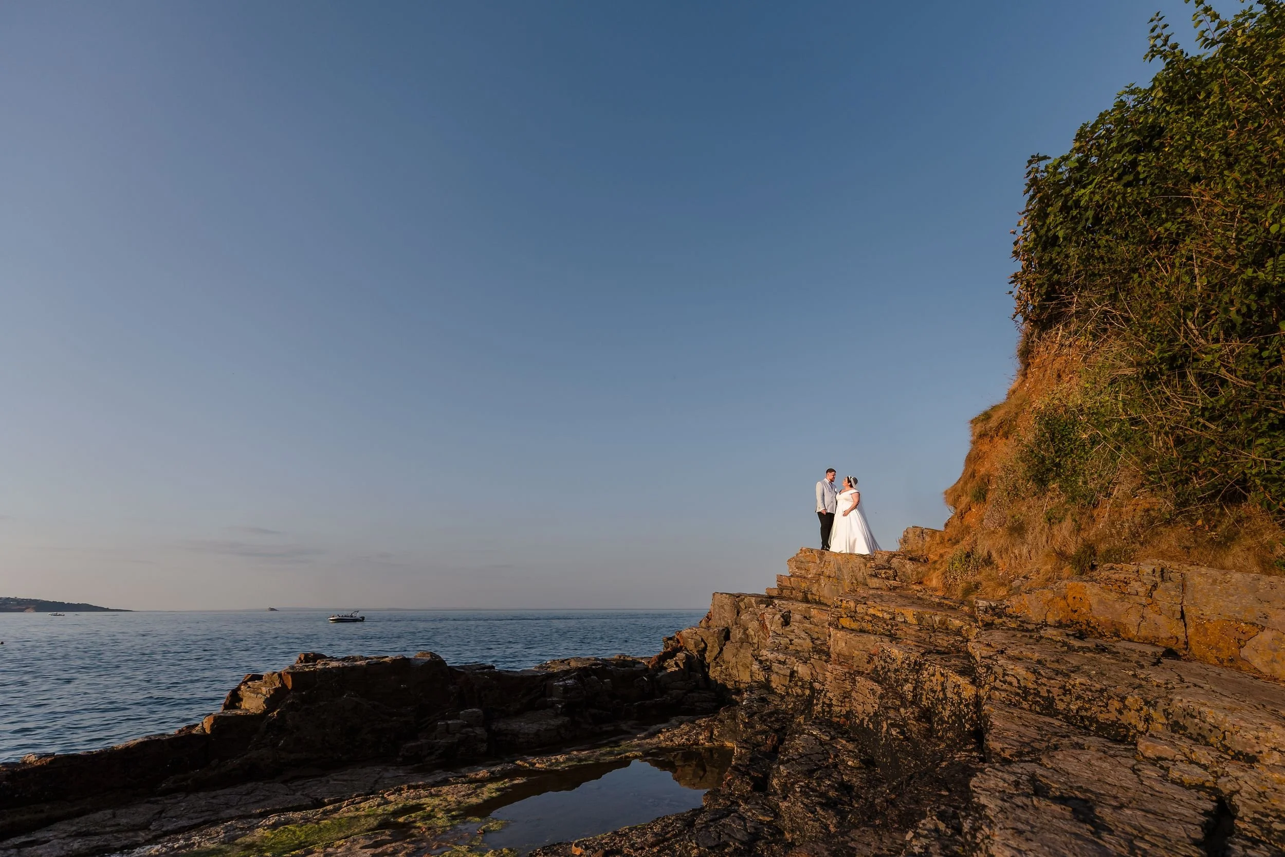 Wide portrait of the bride and groom on the rocks at Berry Head, with a rock pool below and the sea beyond