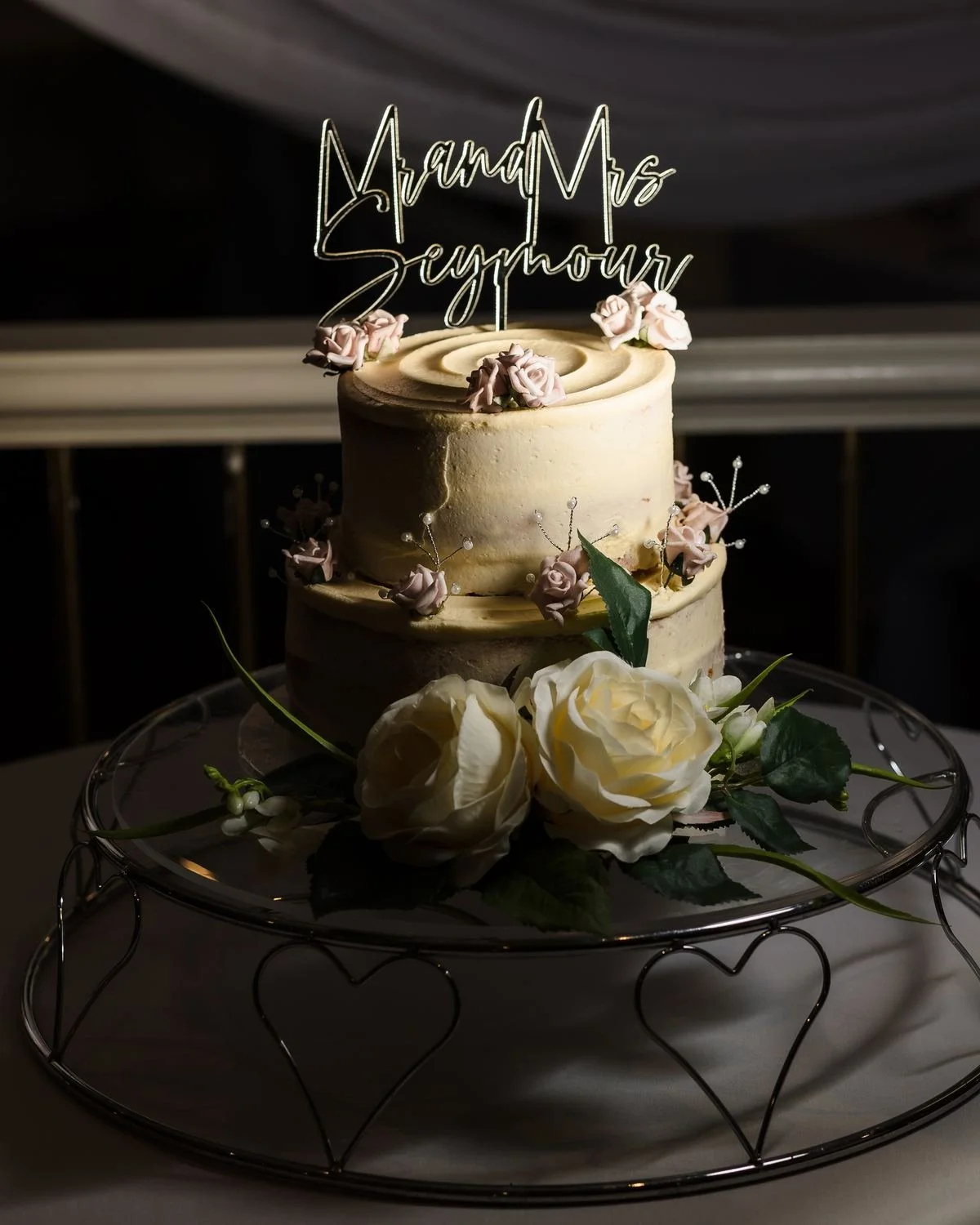 Two-tier white wedding cake with a Mr and Mrs Seymour topper and cream roses, lit with off-camera flash