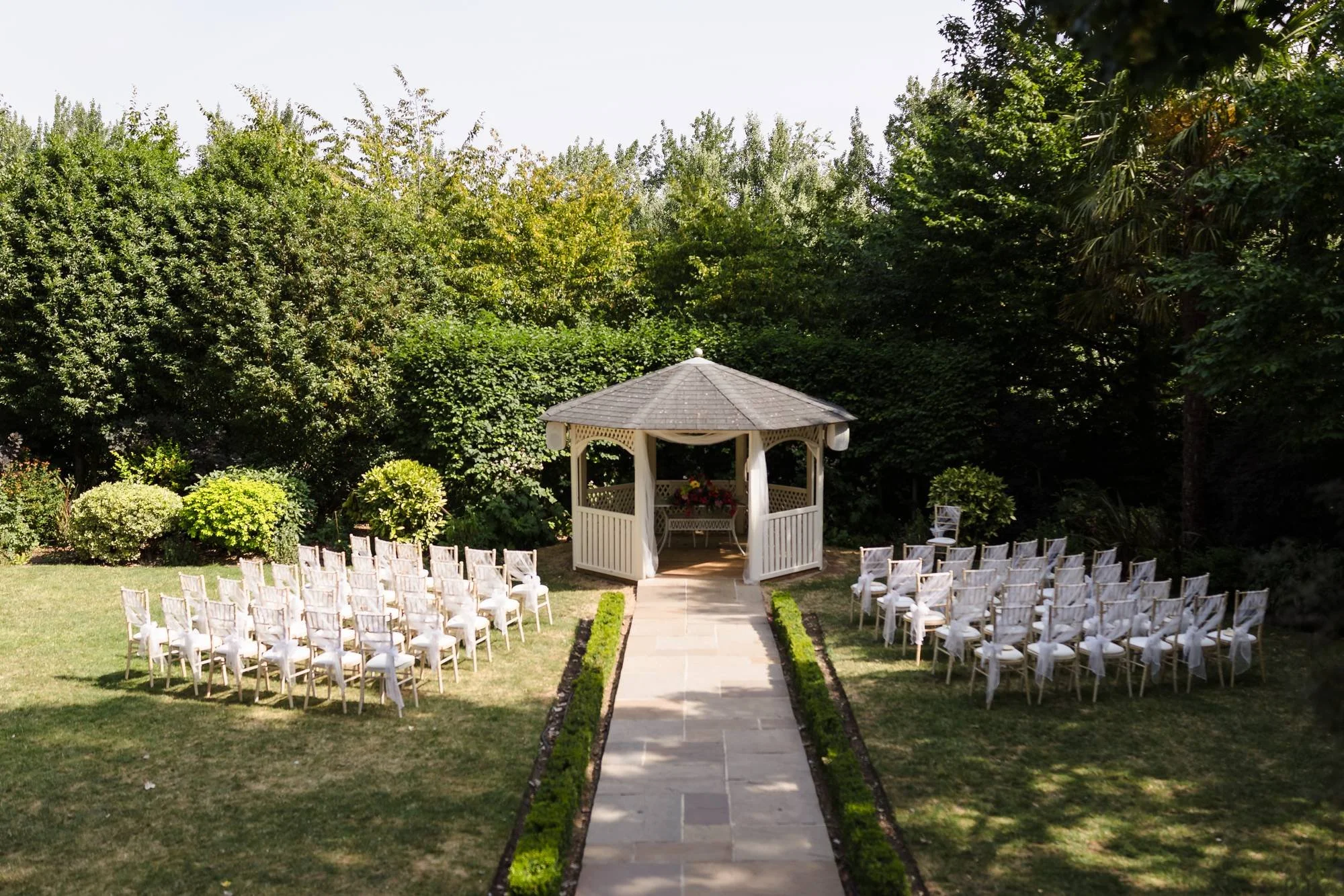 Outdoor ceremony area at Warwick House set up for a summer wedding, with white chairs either side of the aisle to the gazebo