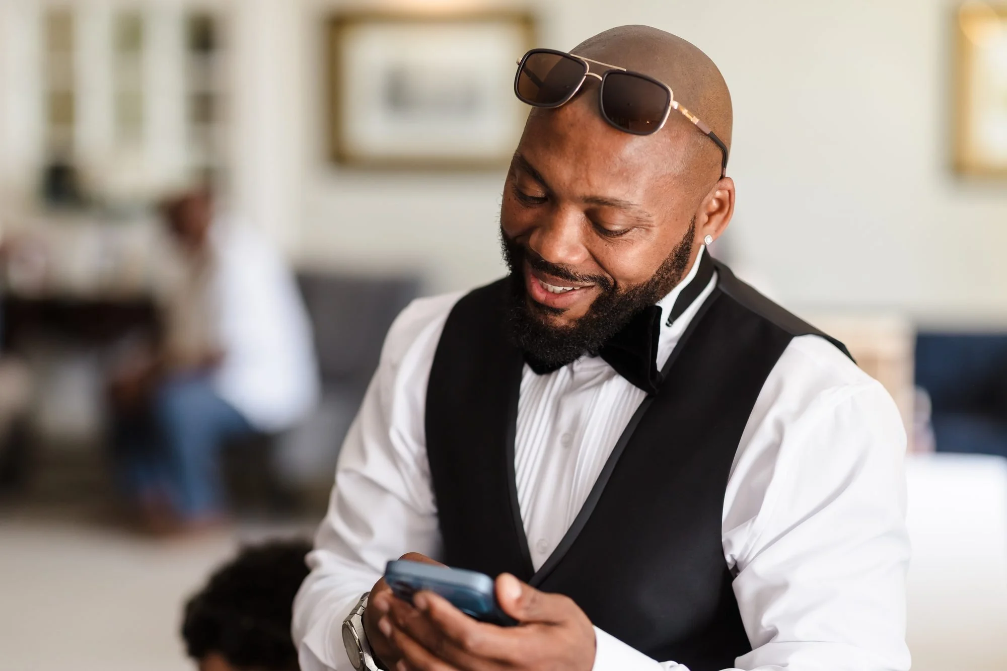 Groom Tafarie smiling at his phone in his waistcoat and bow tie while getting ready at Warwick House