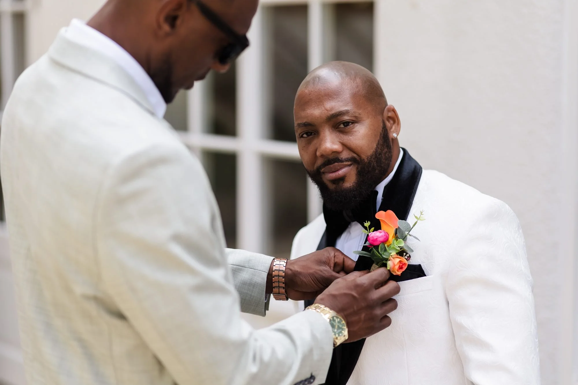 Groomsman pinning a colourful buttonhole on the groom's white tuxedo jacket outside Warwick House