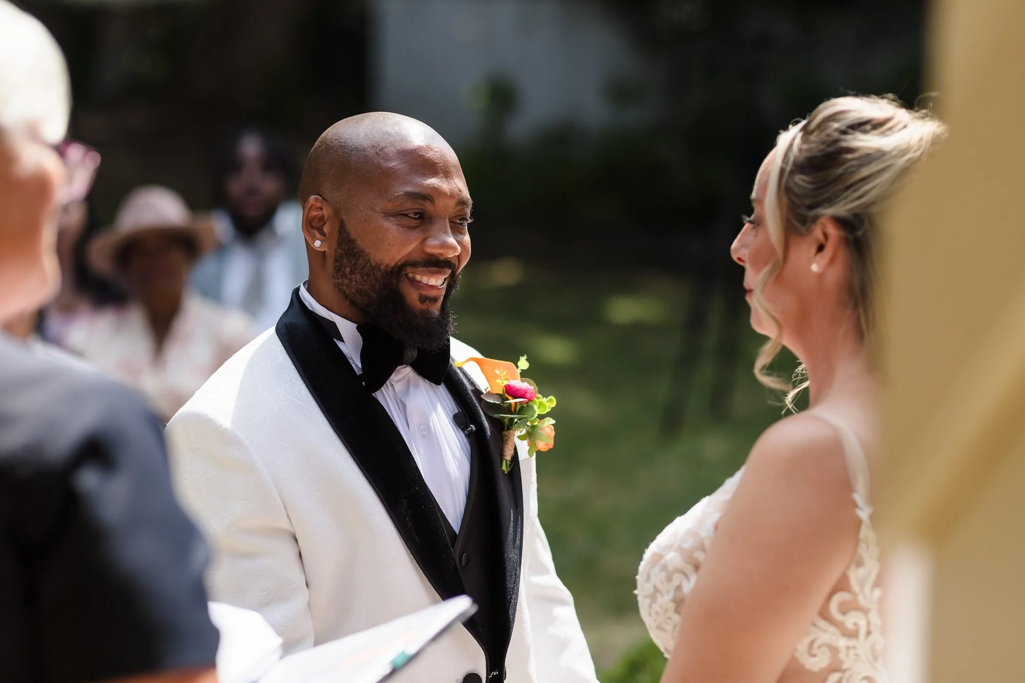 Groom in a white tuxedo smiling at his bride during their outdoor wedding vows at Warwick House