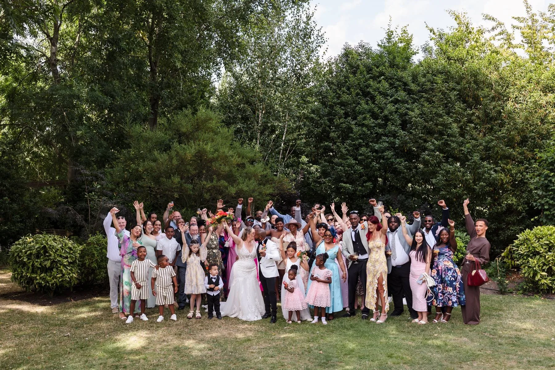 Colourfully dressed wedding guests cheering with arms raised for a big group photo in the gardens at Warwick House