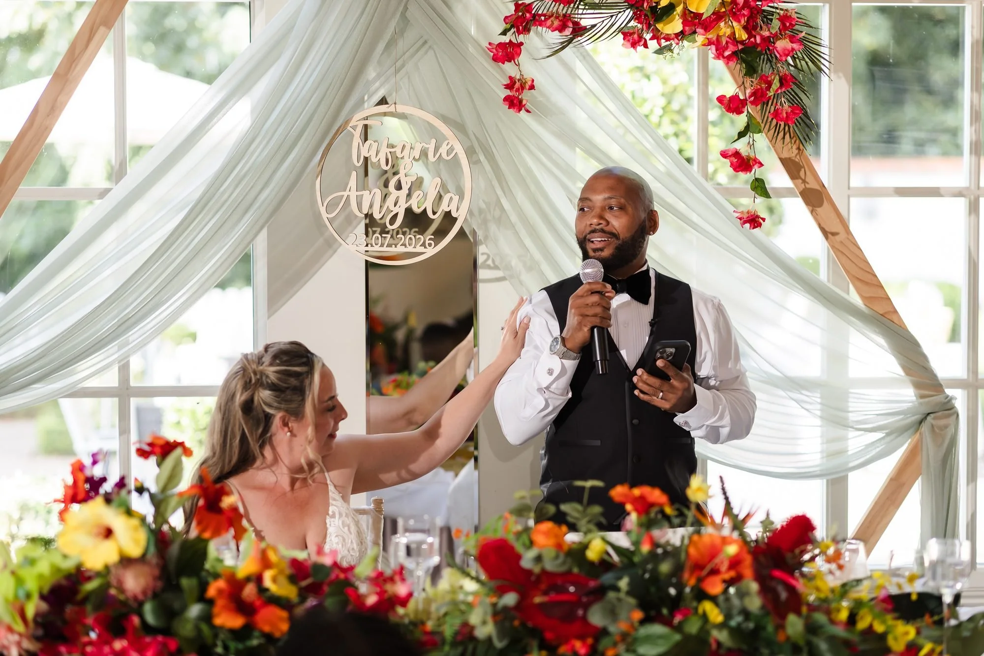 Groom giving his speech at the top table as the bride reaches up to him, under a flower arch at Warwick House