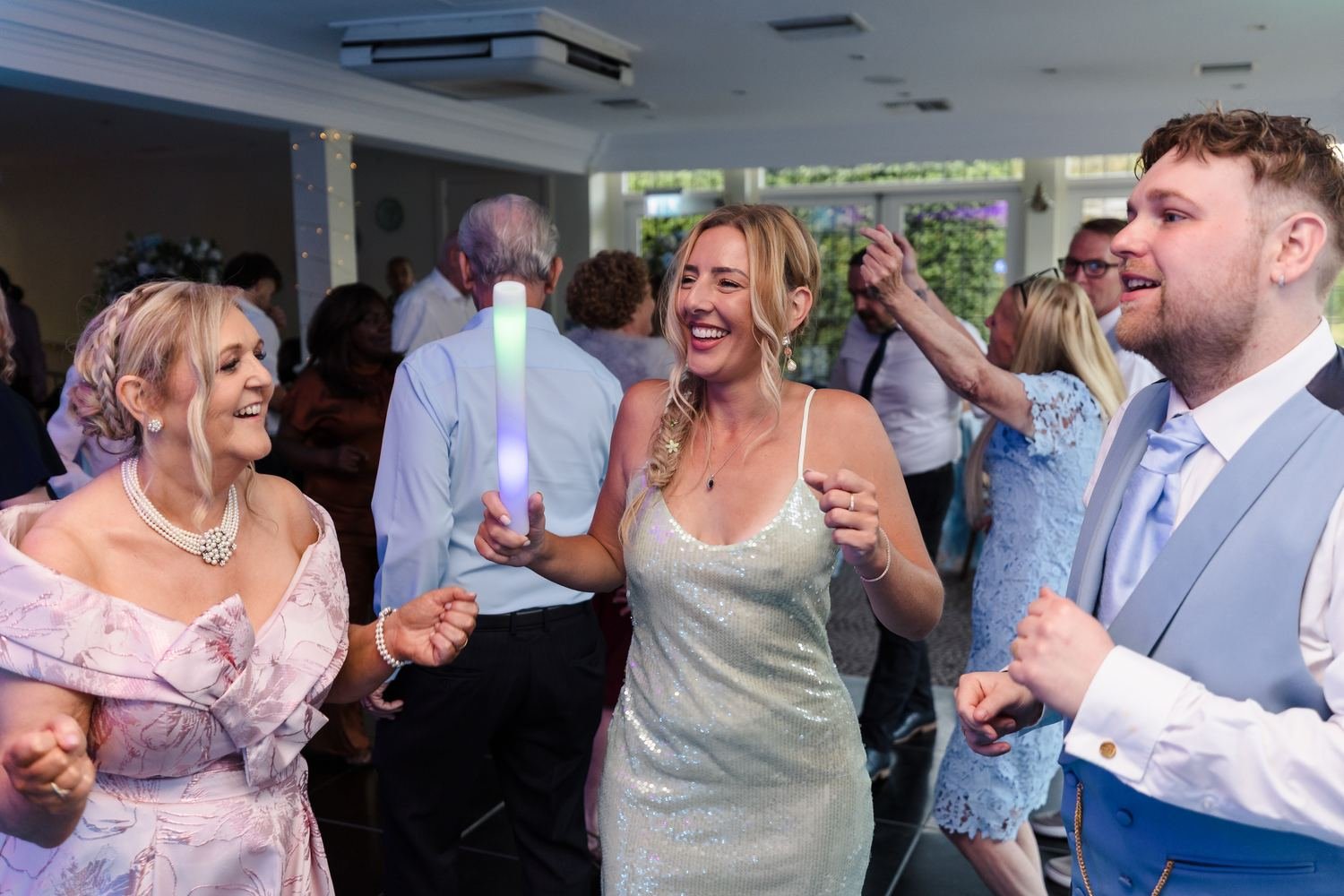 Guests laughing and dancing with a light-up baton alongside one of the grooms on the dance floor