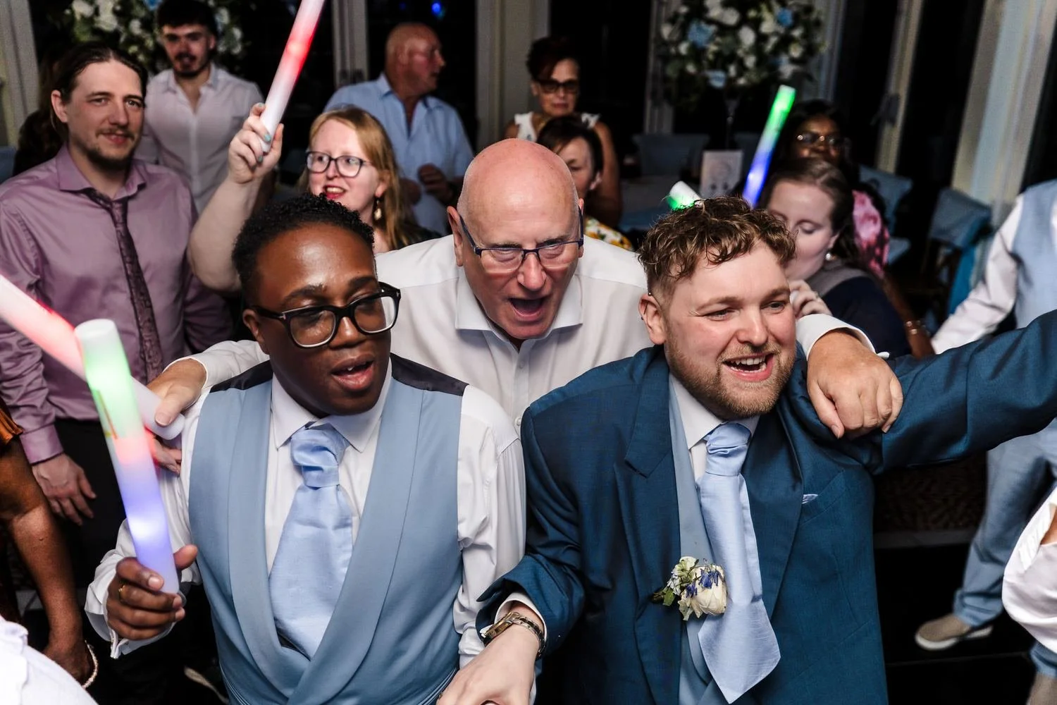 Liam and Shaun in the middle of the dance floor, arms around a guest, surrounded by friends waving light-up batons