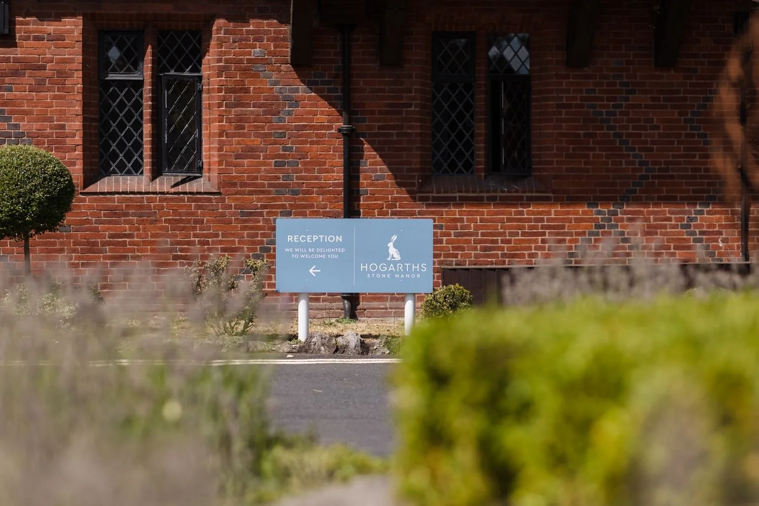 Blue Hogarths Stone Manor reception sign in front of the red-brick building, framed by soft foreground planting