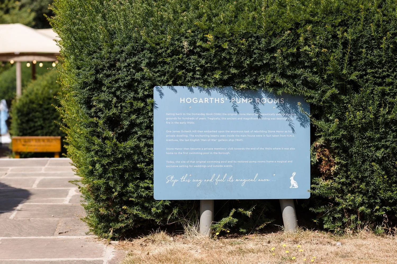 Hogarths' Pump Rooms history sign set into a tall yew hedge beside the path to the outdoor ceremony area