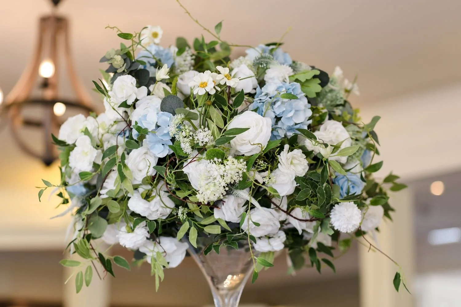 Tall table centrepiece of white roses, pale blue hydrangeas, daisies and trailing greenery in a glass vase