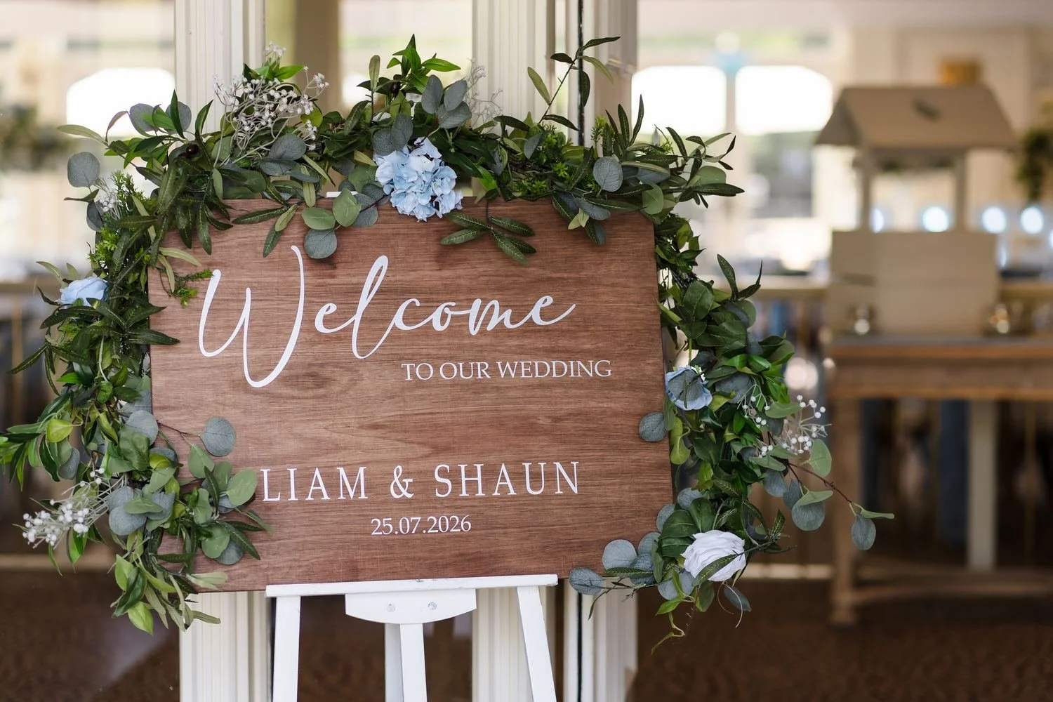 Wooden welcome sign reading Liam and Shaun 25.07.2026, trimmed with eucalyptus, greenery and pale blue hydrangea