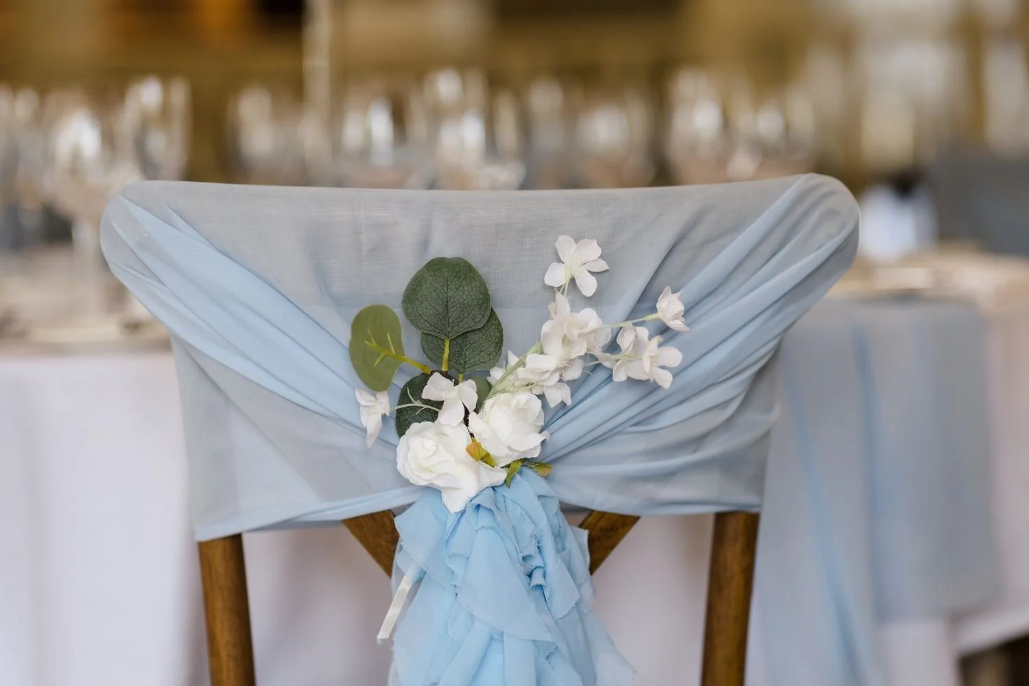 Close-up of a pale blue chiffon chair sash finished with white flowers and eucalyptus on a wooden cross-back chair