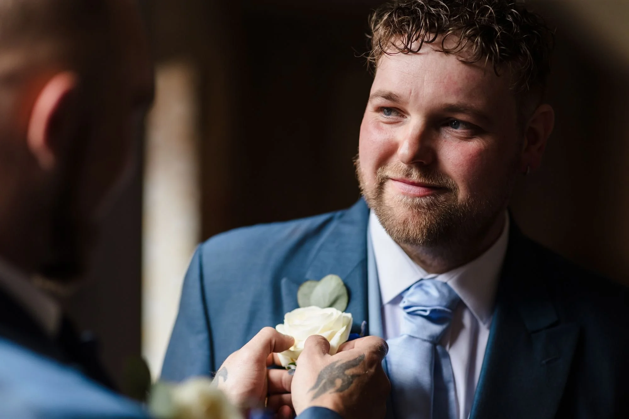 One of the grooms smiling as a white rose buttonhole is pinned to his blue jacket